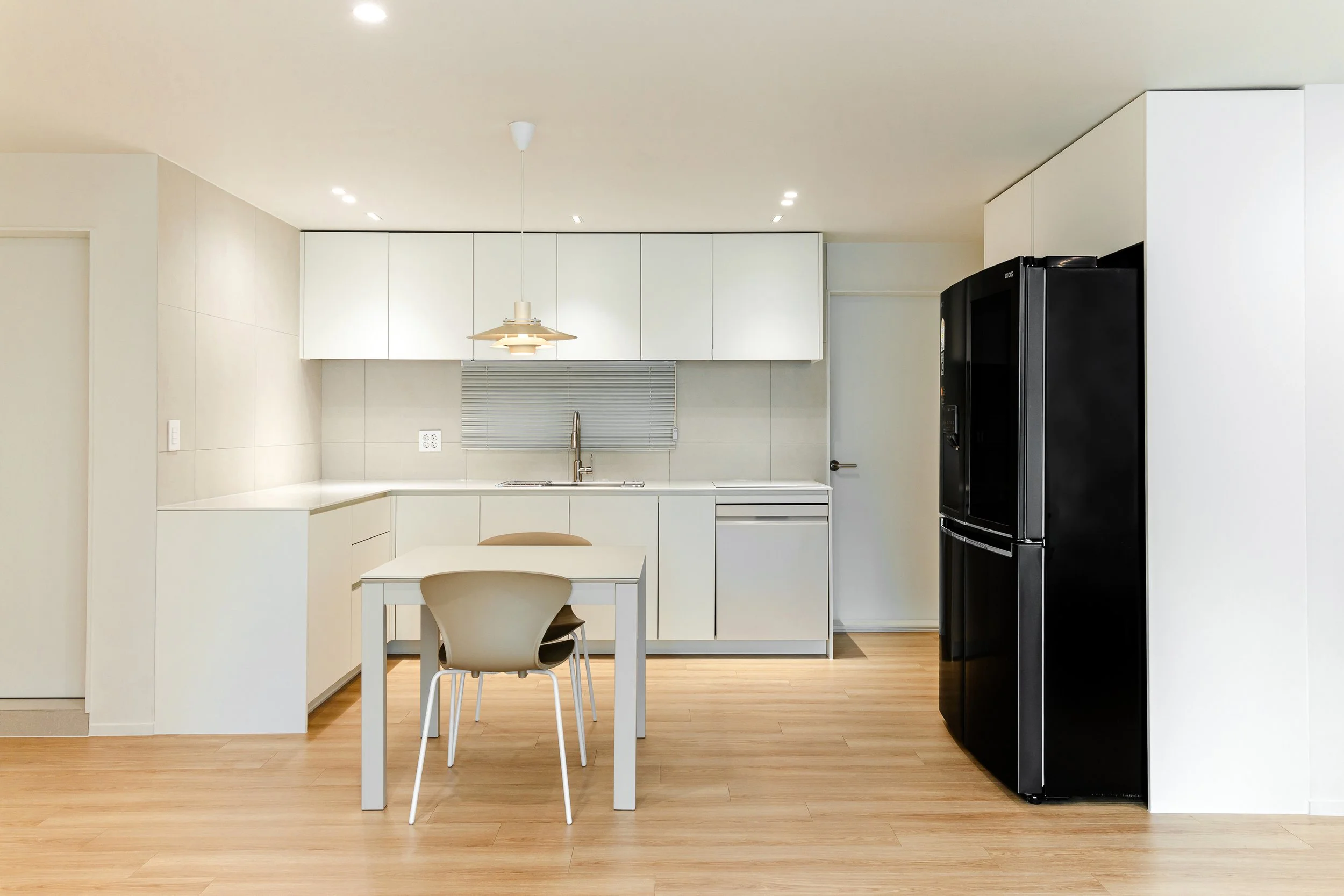 Modern minimalist kitchen with white cabinets, a black refrigerator, light wooden floors, a small dining table with two chairs, and a window with blinds above the sink.