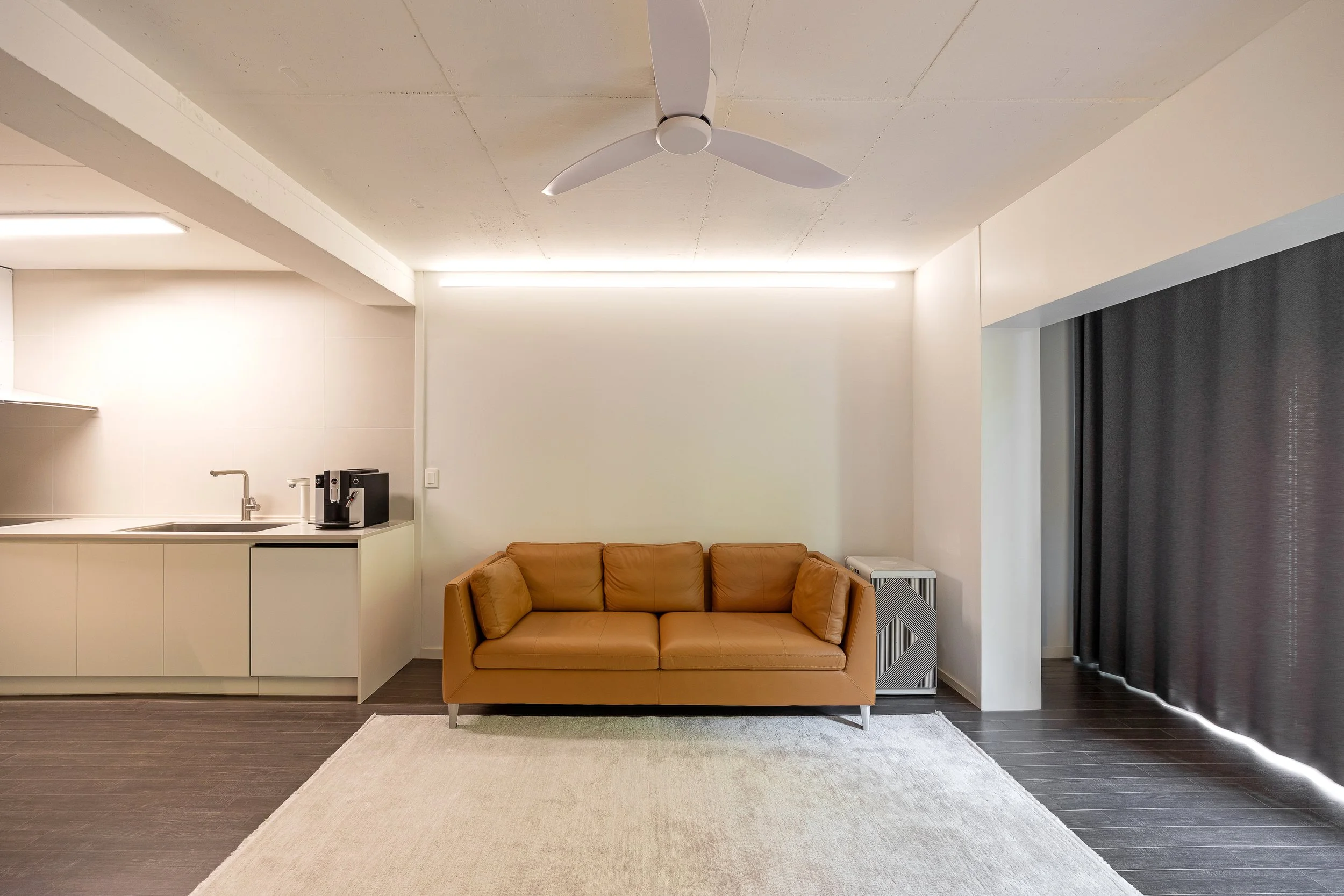 Modern living room with a tan leather sofa, white walls, dark wood floor, and sliding dark curtains on the right. A large white rug is in front of the sofa, and a white ceiling fan is overhead. The kitchen area with a sink and coffee maker is visible