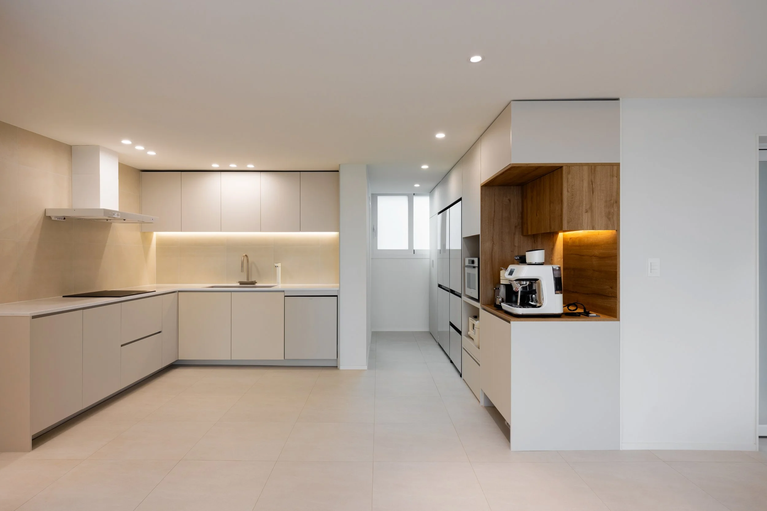 Modern kitchen with minimalist white cabinets, beige backsplash, built-in appliances, and a coffee machine on a wooden shelf.