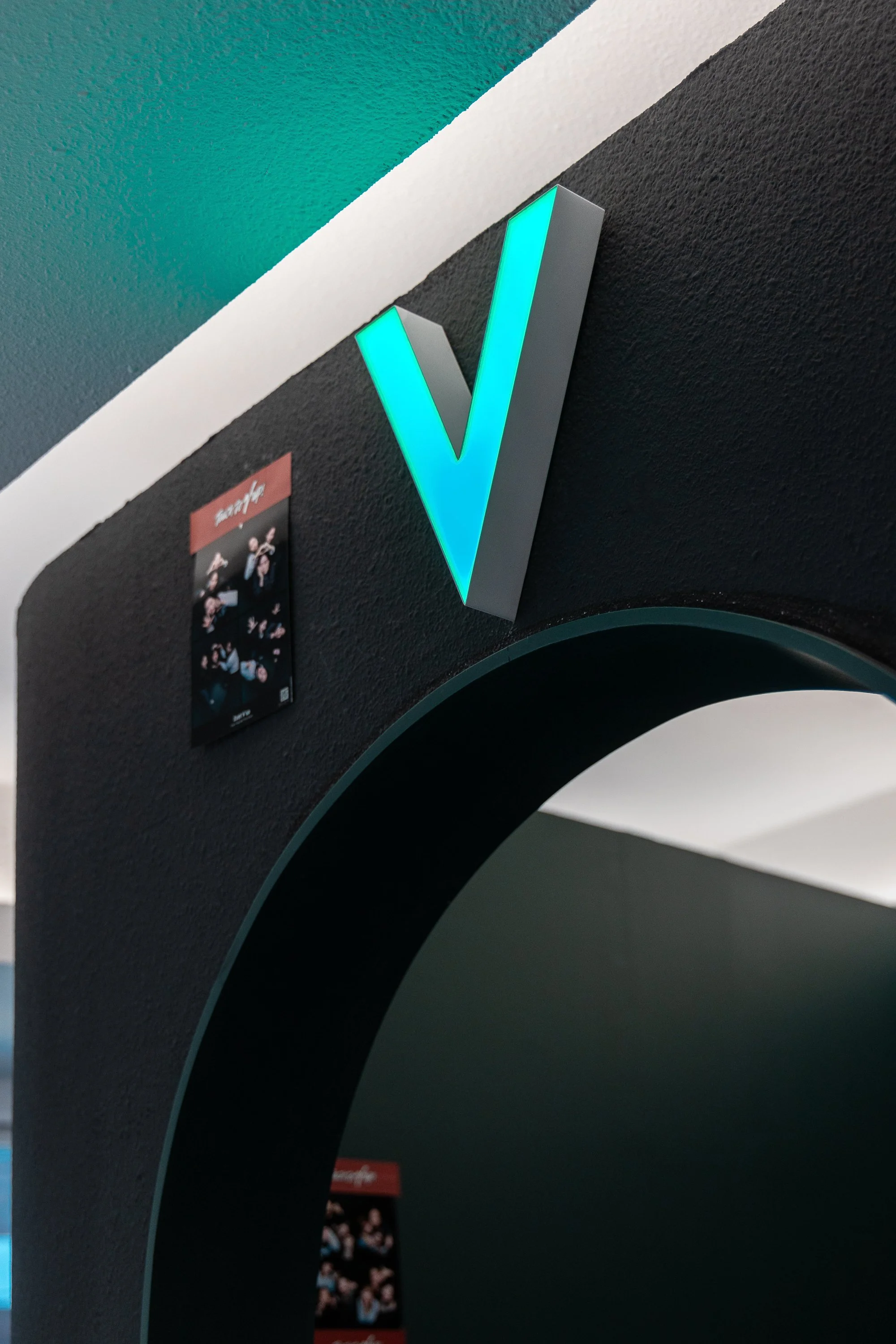A large, illuminated teal letter V mounted on a black textured wall inside a modern space.