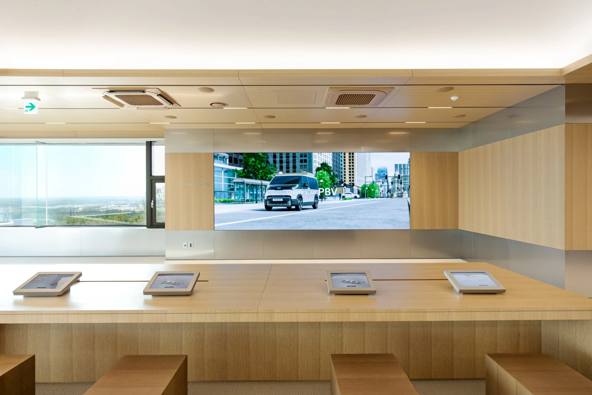 A modern interior space with a wooden counter and four digital screens, a large window revealing a cityscape, and a large digital display showing a vehicle and city scene.