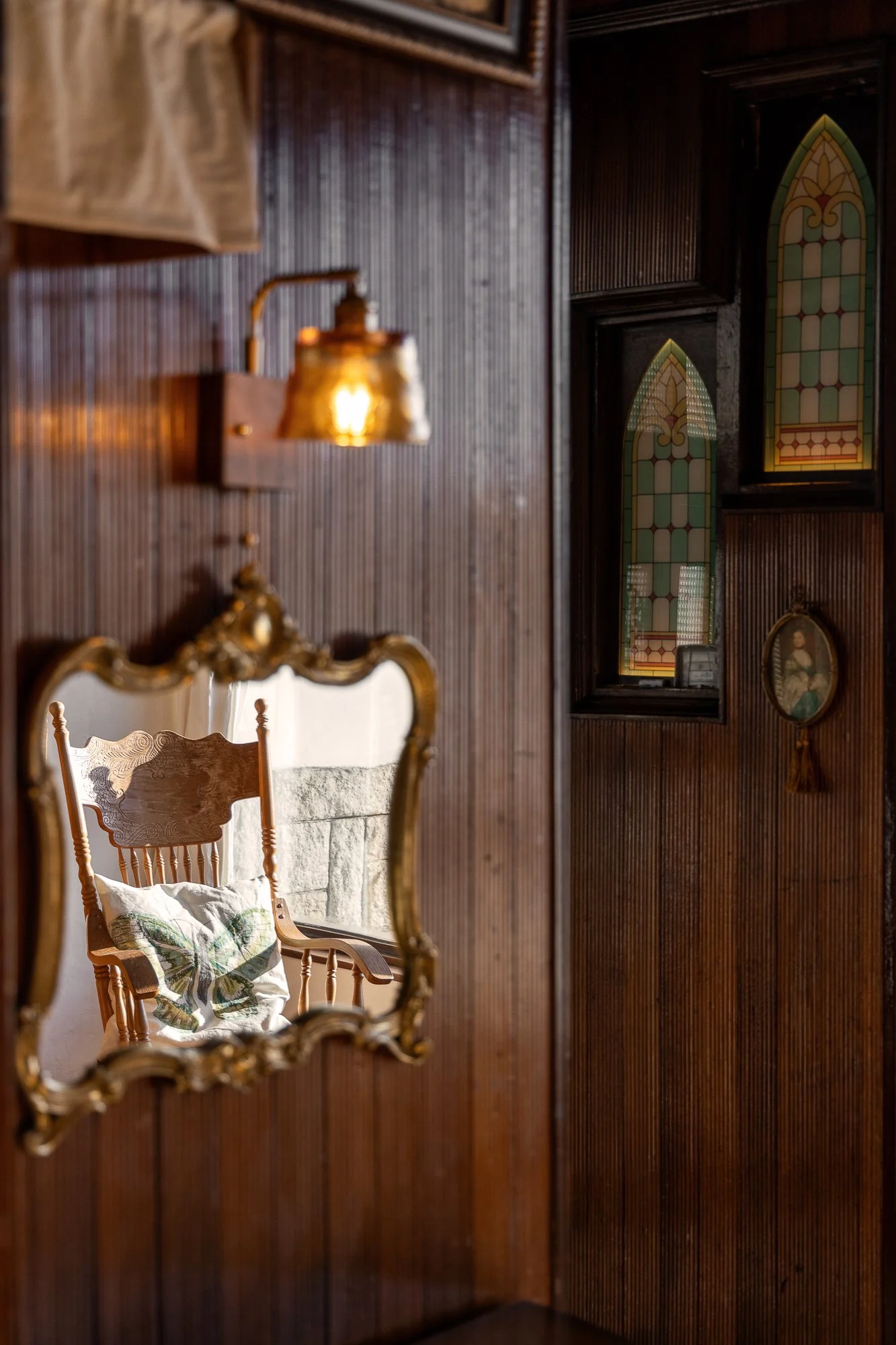 A reflection of a wooden rocking chair with a floral cushion in a vintage ornate mirror hanging on a wooden-paneled wall. The chair is next to a stone wall, and there are small stained glass windows and a framed portrait on the wall.