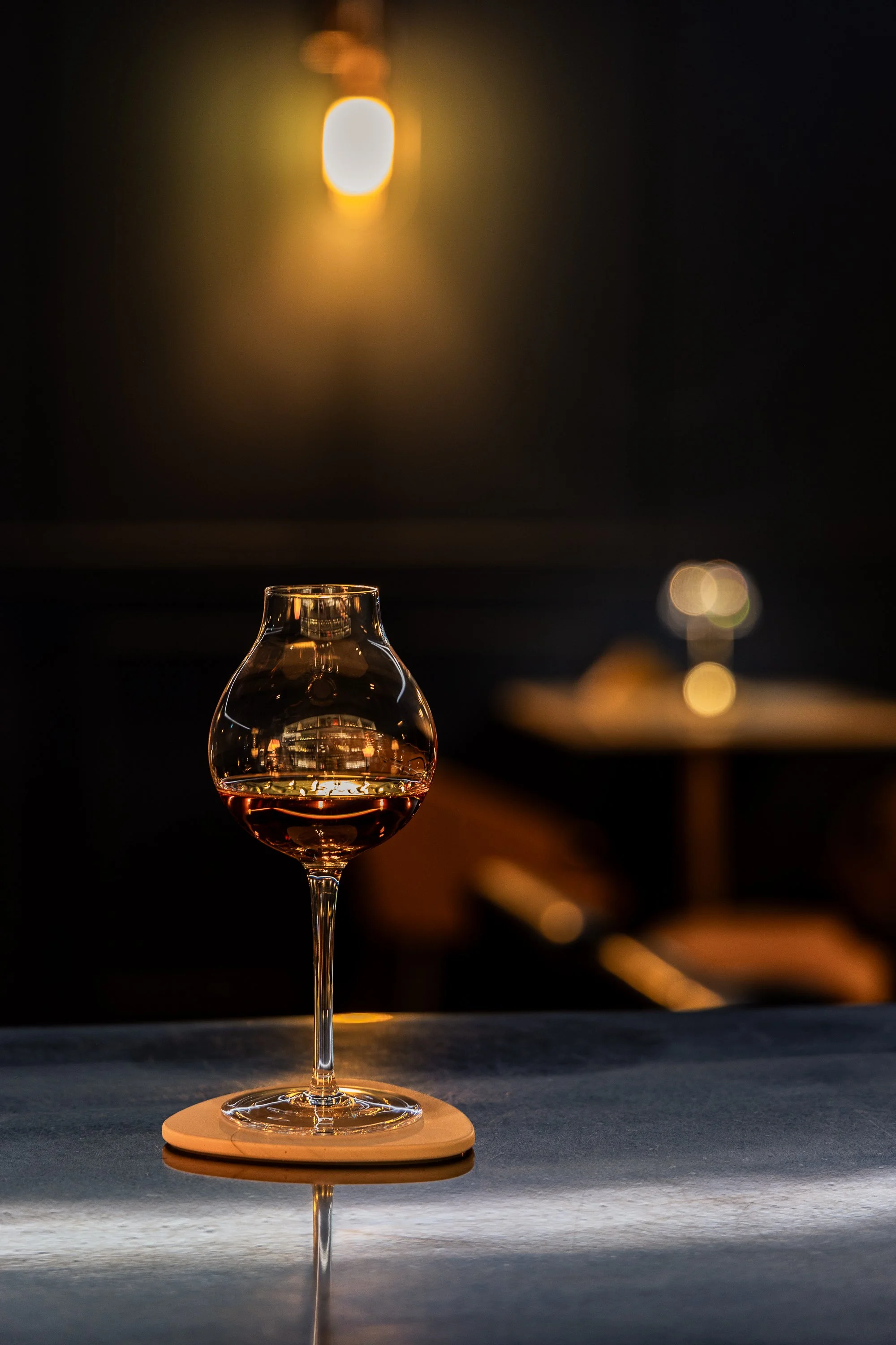 A glass of whiskey on a coaster, placed on a dark bar counter, with warm ambient lighting reflecting off the glass.