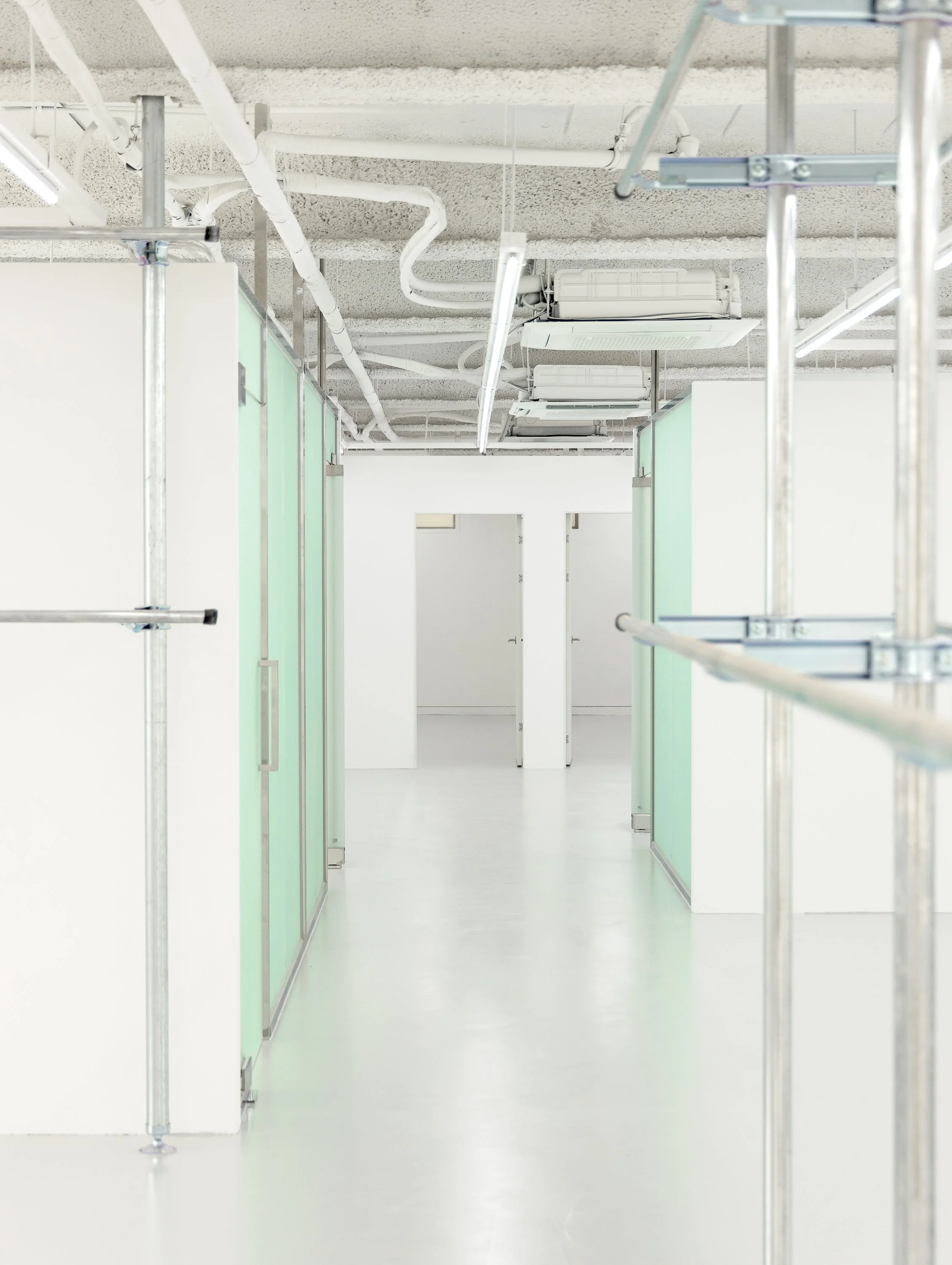 Empty office or studio space with white walls, light-colored flooring, and partitions with frosted glass panels; ceiling with exposed pipes and ducts, fluorescent lighting, and air conditioning units.