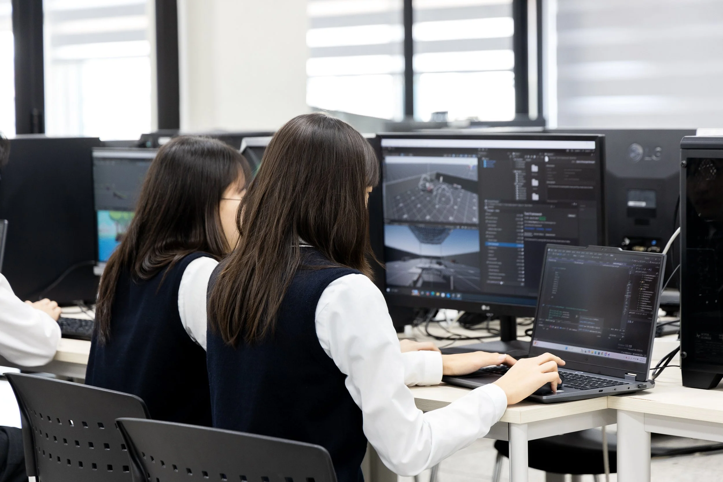 Two students working on computers, one using a laptop and the other using a desktop, in a classroom or lab setting.