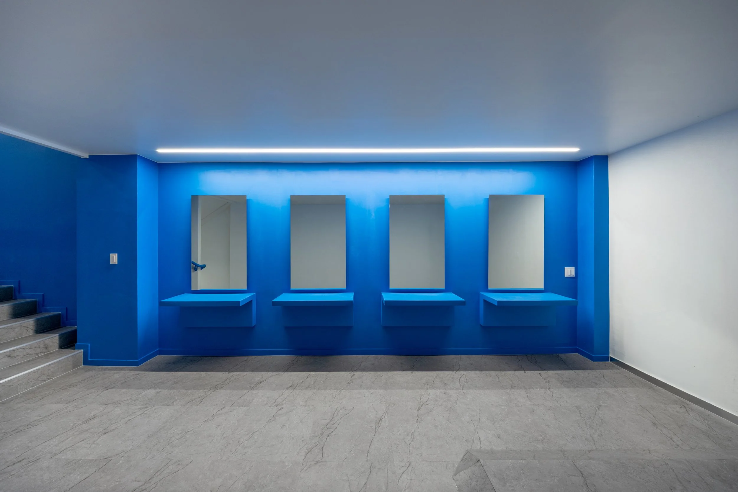 Interior of a modern space with four rectangular mirrors on a bright blue wall, each mirror has a blue ledge below it, with a staircase visible on the left, and the floor is light gray with subtle marbling.