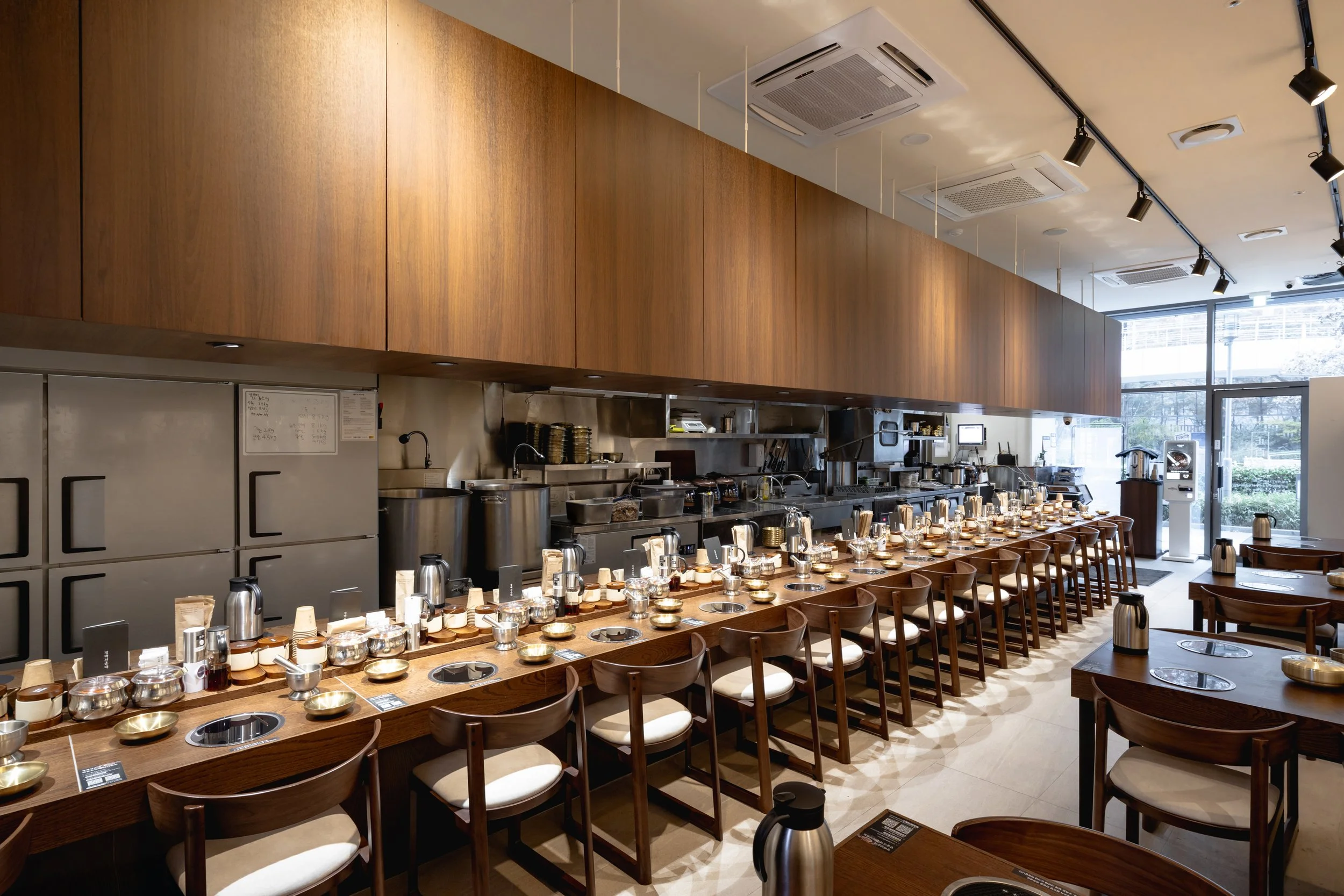 Interior of a restaurant with a long wooden counter setup with multiple dining chairs, teapots, and utensils, with an open kitchen area and large windows letting in natural light.