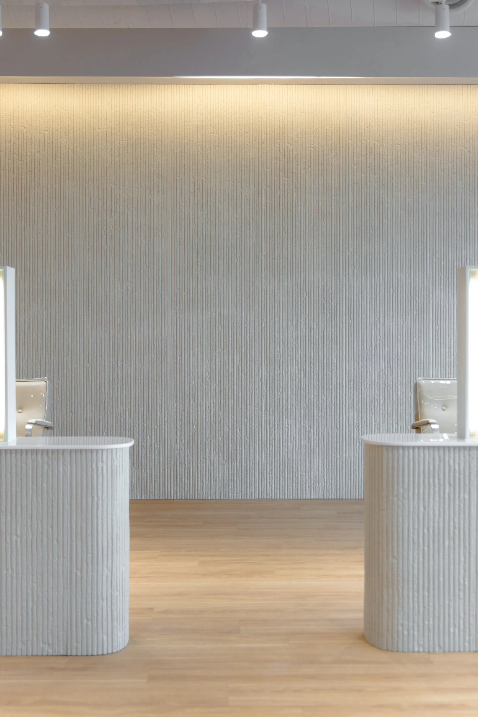 Two white reception desks with mirrors and chairs in a minimalist, modern lobby with textured white walls and wood flooring.