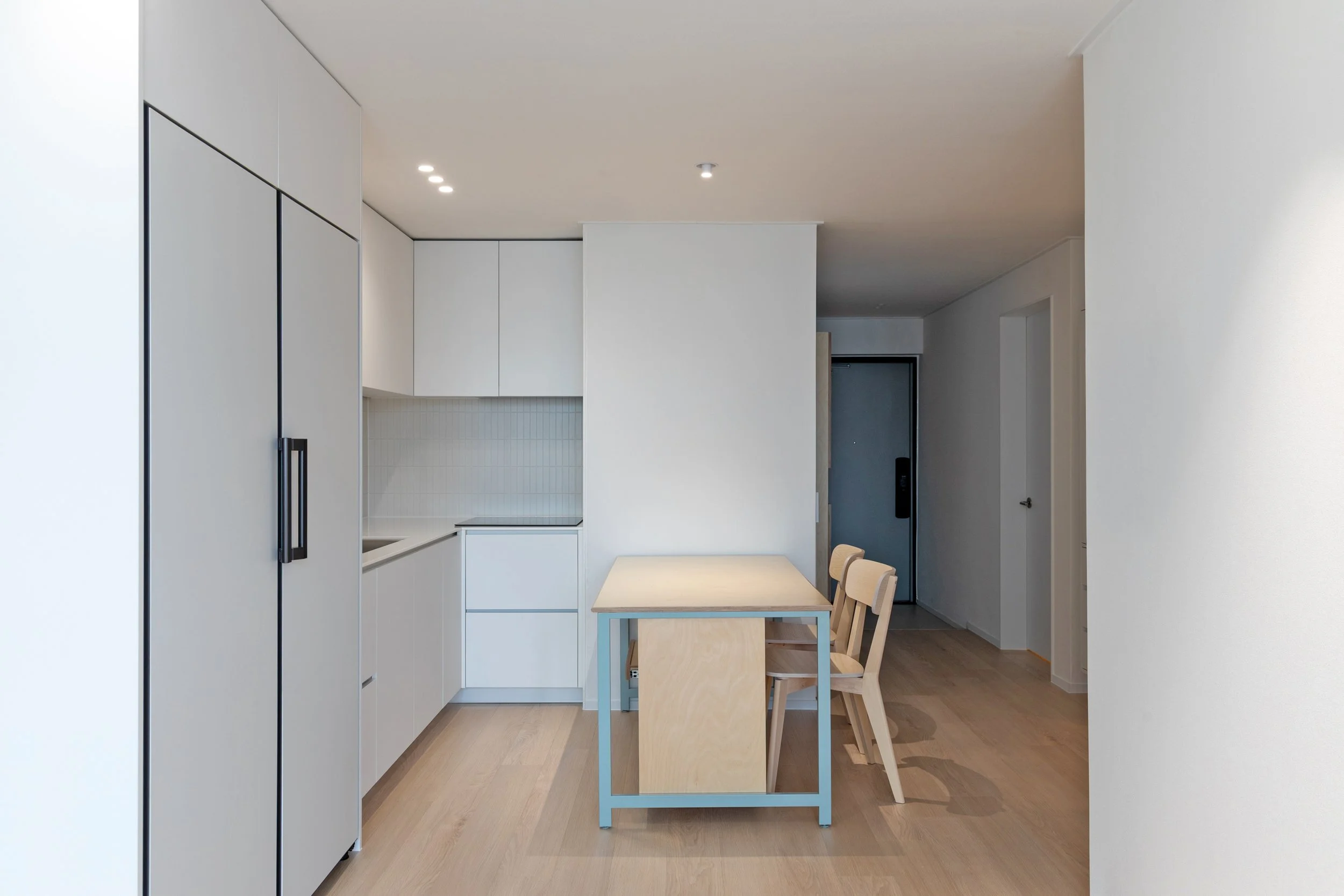 Minimalist kitchen and dining area with light wood flooring, white cabinets, a small table with a light wood top, and three matching light wood chairs.