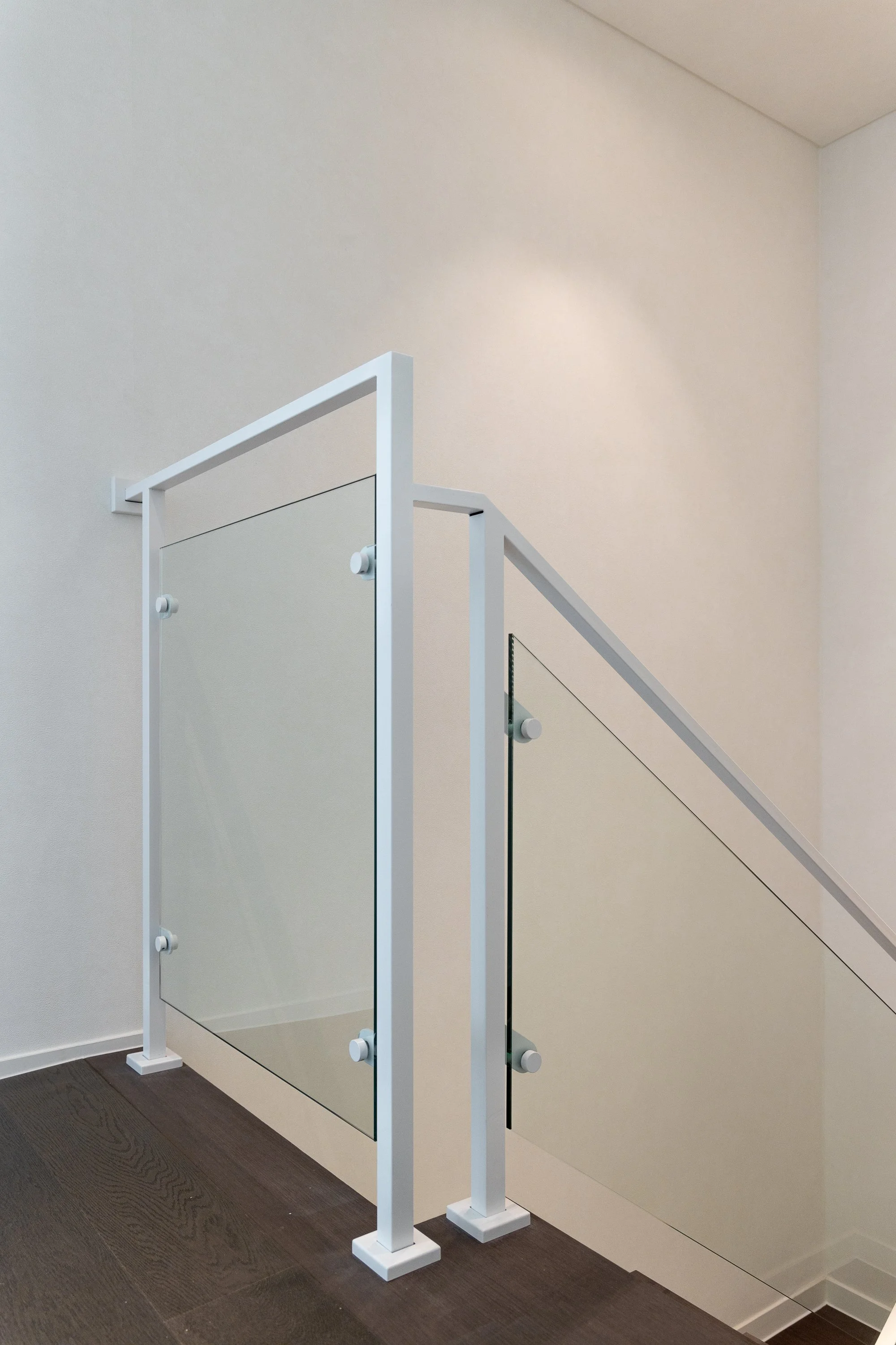 Modern staircase railing with a glass panel, white frame, and rectangular base, located inside a building with light-colored walls and dark wooden flooring.