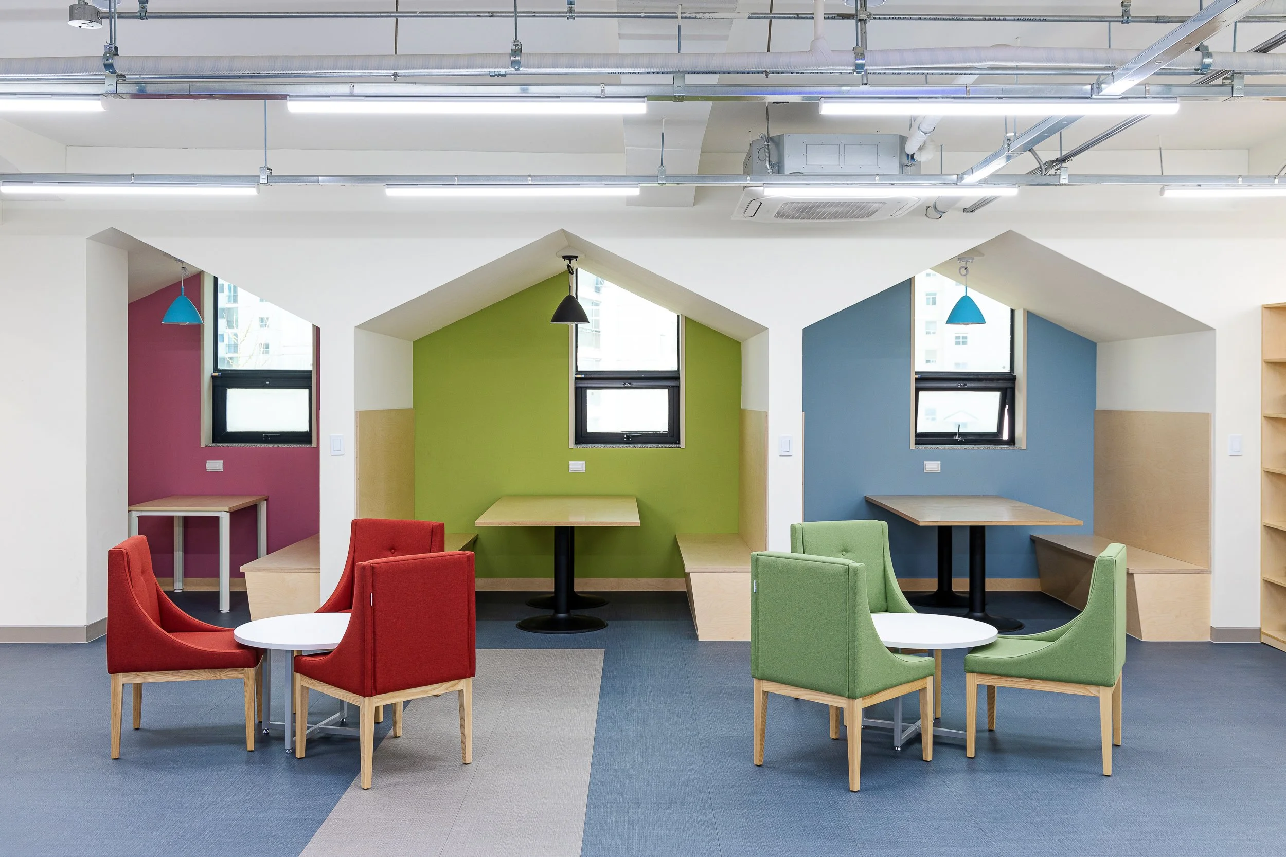 Colorful modern seating area with red and green chairs, small tables, and cozy alcoves with colorful walls and windows in a well-lit room.