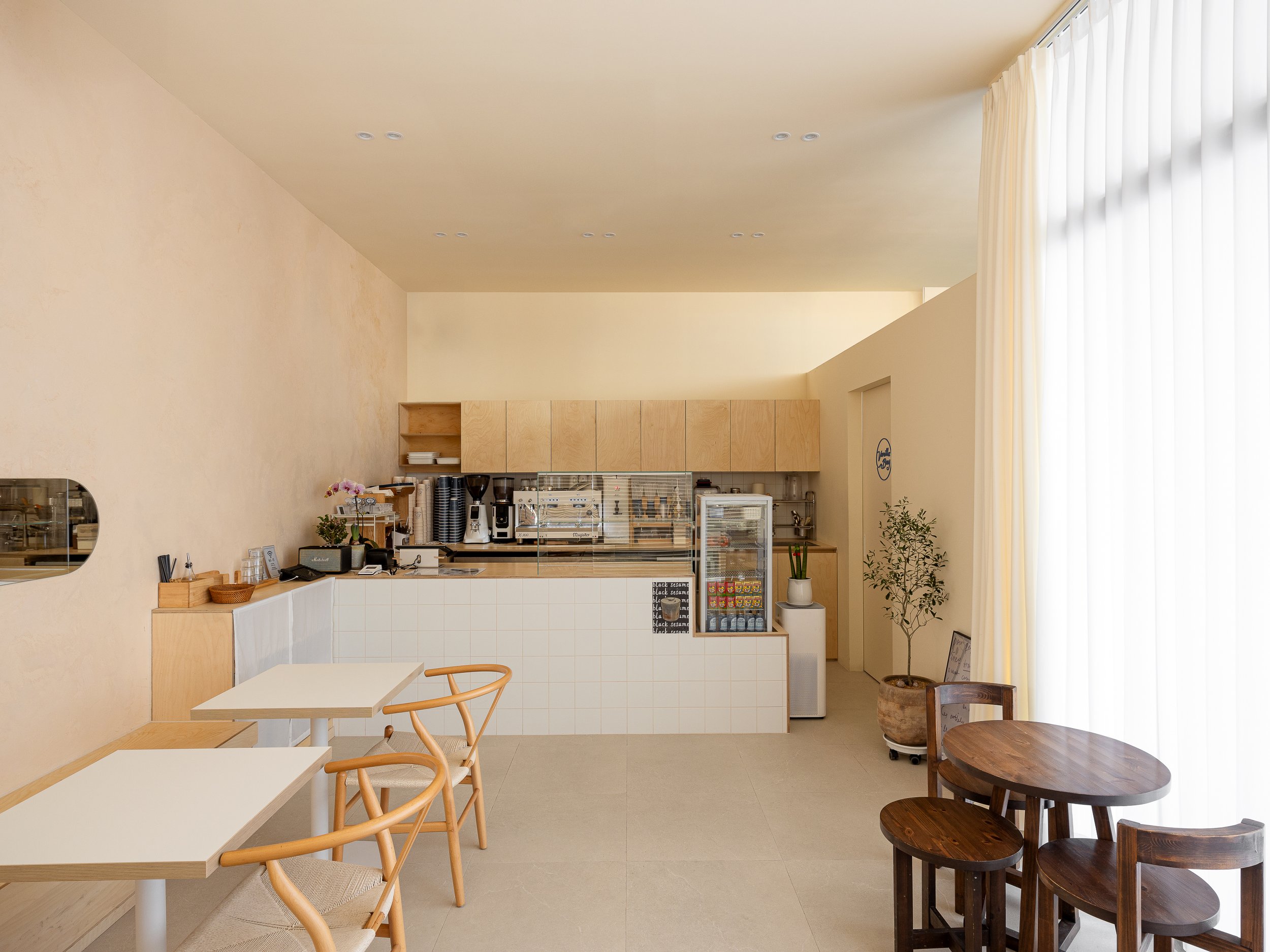 Minimalist cafe with wooden tables and chairs, coffee machines, a fridge, potted plants, and large curtains on windows.
