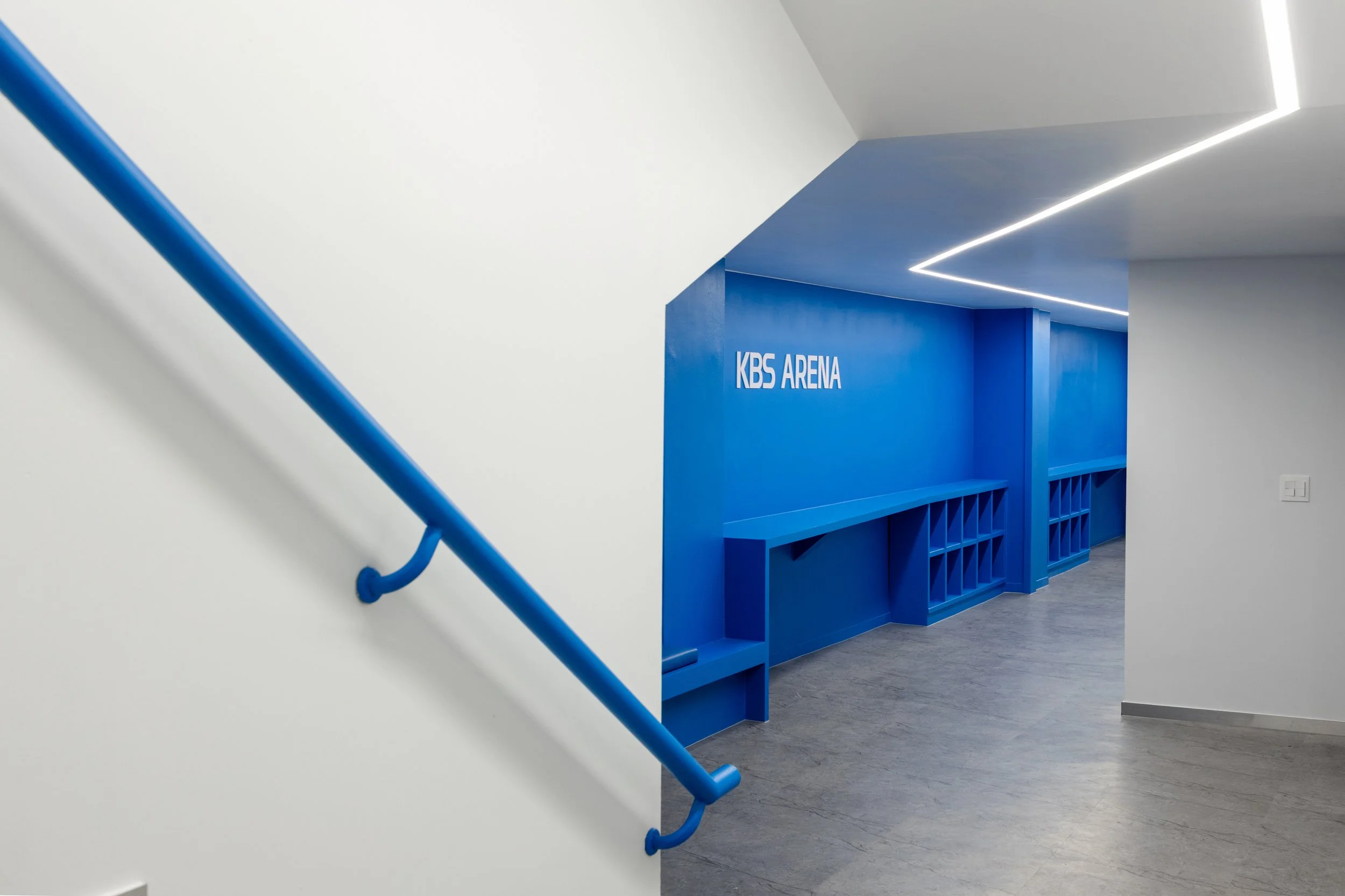 Modern interior of KBS Arena with a white wall, blue handrail, and blue wall with shelving units.