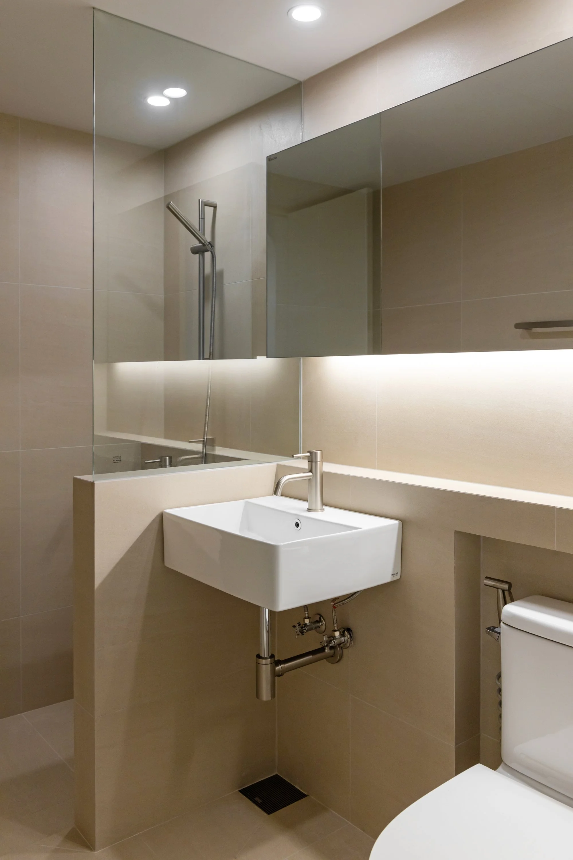 A modern bathroom featuring a white rectangular sink with a sleek faucet, above a beige tiled wall. A large mirror with indirect lighting is mounted behind the sink. To the right, part of a white toilet is visible. A reflection in the mirror shows a 
