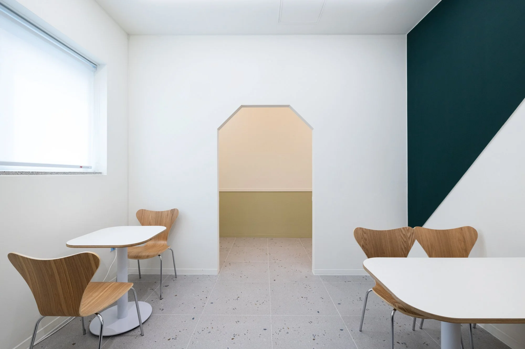 A small, modern room with white and dark green walls, a large window, and minimalist wooden chairs and white tables.