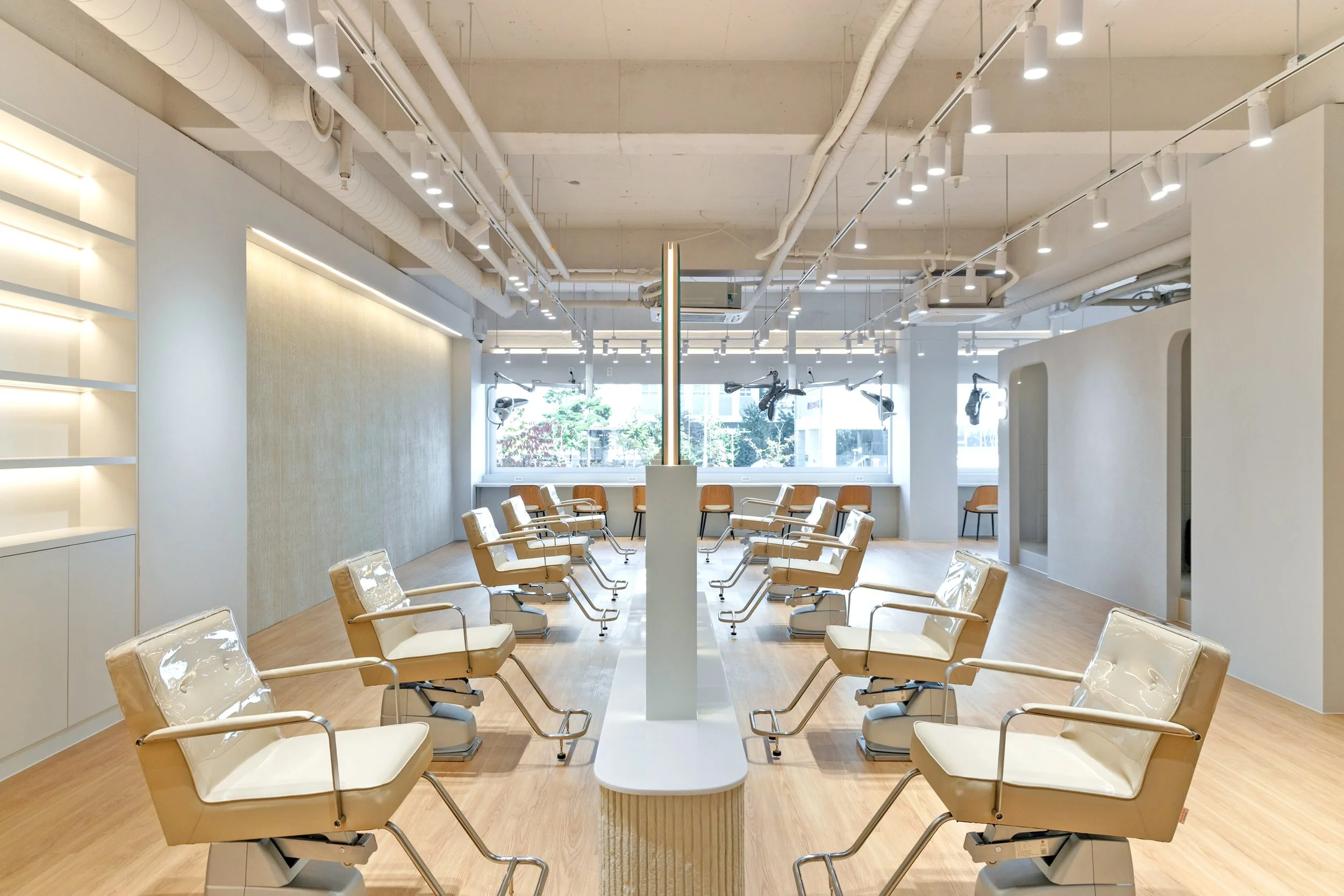 Empty salon chairs in a modern hair salon with large windows, white walls, wooden flooring, and overhead lighting.