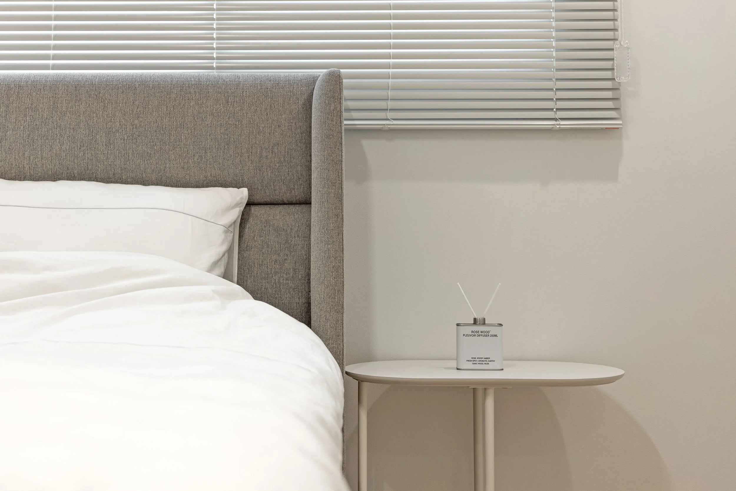 A bedroom corner with a bed, grey upholstered headboard, white bedding, a white side table, and a small diffuser on the table, situated near window blinds.
