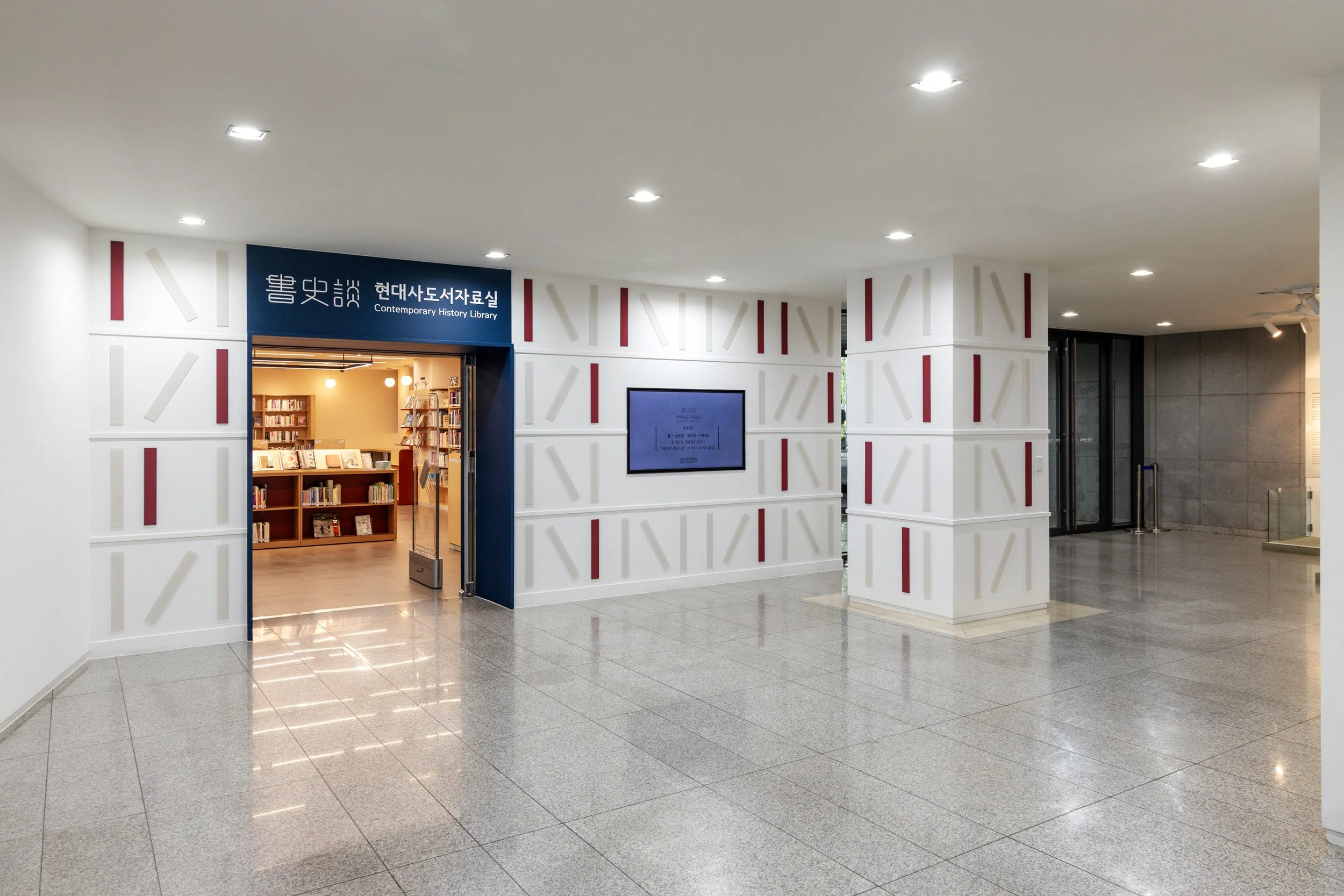 Interior view of a modern library entrance with a sign that reads 'Contemporary History Library' in Korean and English, featuring white walls with red and beige geometric patterns, a digital display on the wall, and a glimpse of wooden bookshelves in