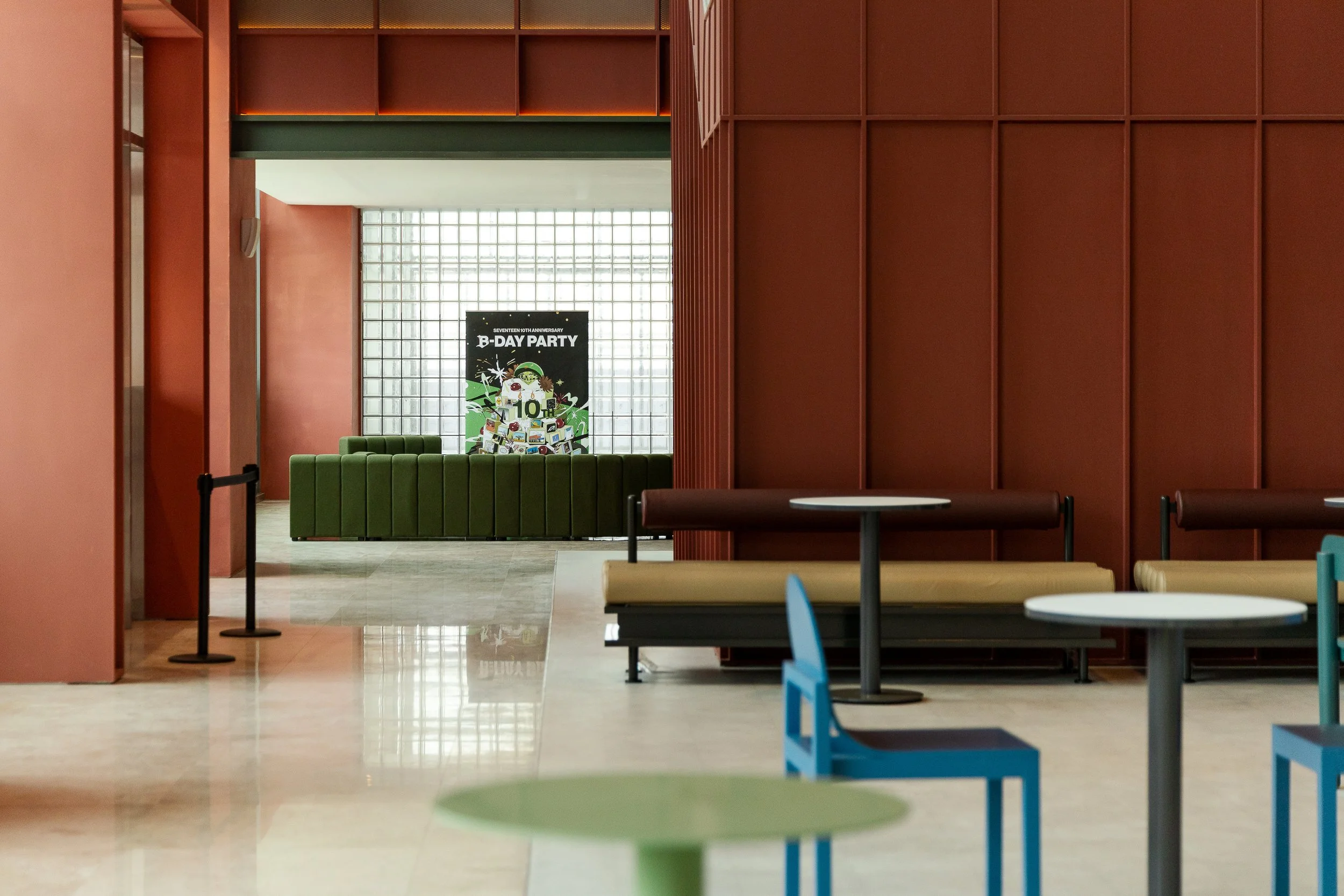 Interior of a modern space with red walls, round tables, and colorful chairs. In the background, there's a green sofa and a large window with a grid pattern. A poster with green, black, and white colors celebrating a 10th birthday is visible.