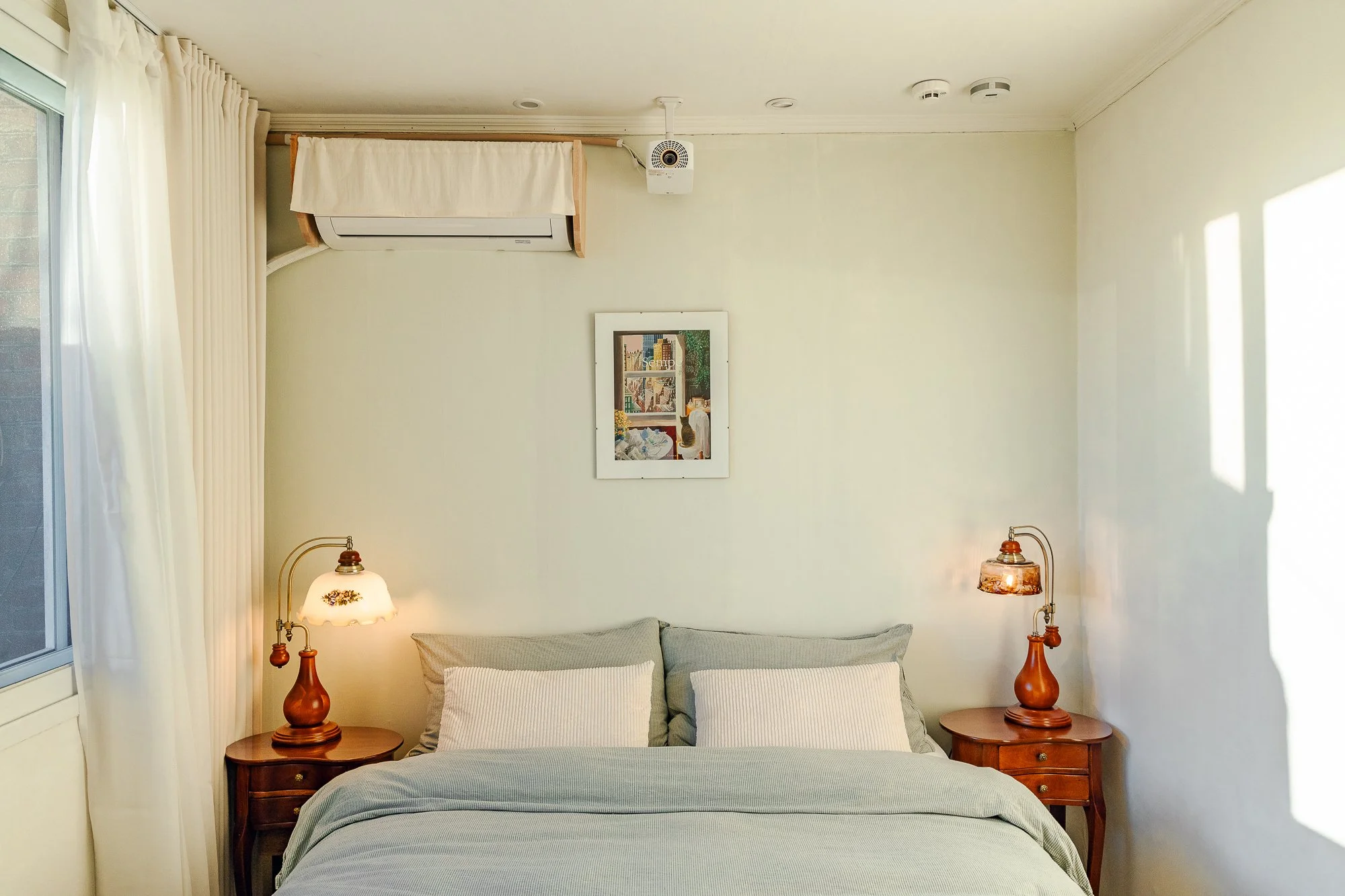 A neatly made bed with gray bedding and two white pillows, flanked by two wooden nightstands with vintage lamps, beside a window with white curtains, in a cozy bedroom.