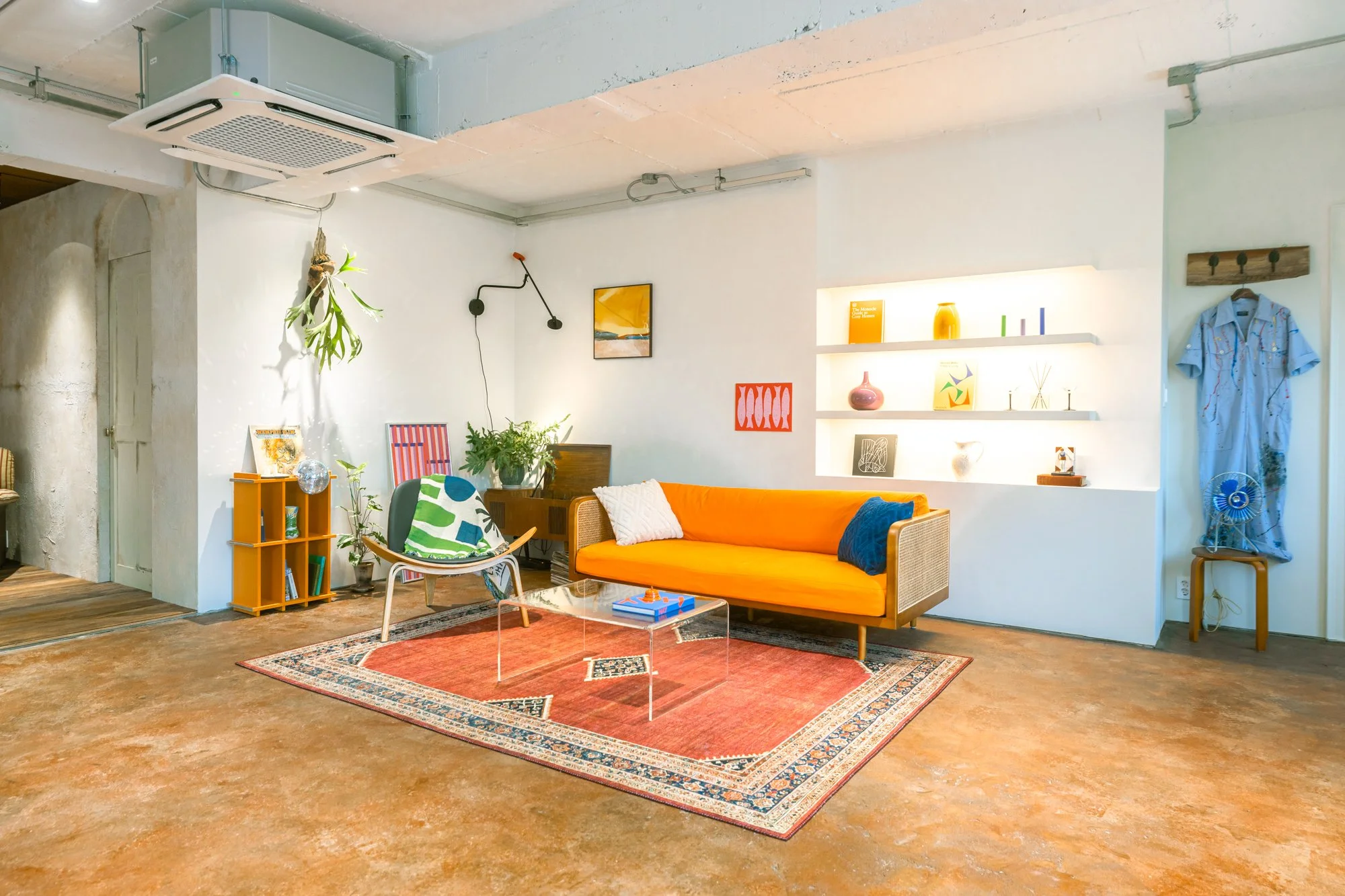 Modern living room with a vibrant orange sofa, colorful patterned rug, and eclectic decor, including plants, artwork, and shelving with various decorative items.