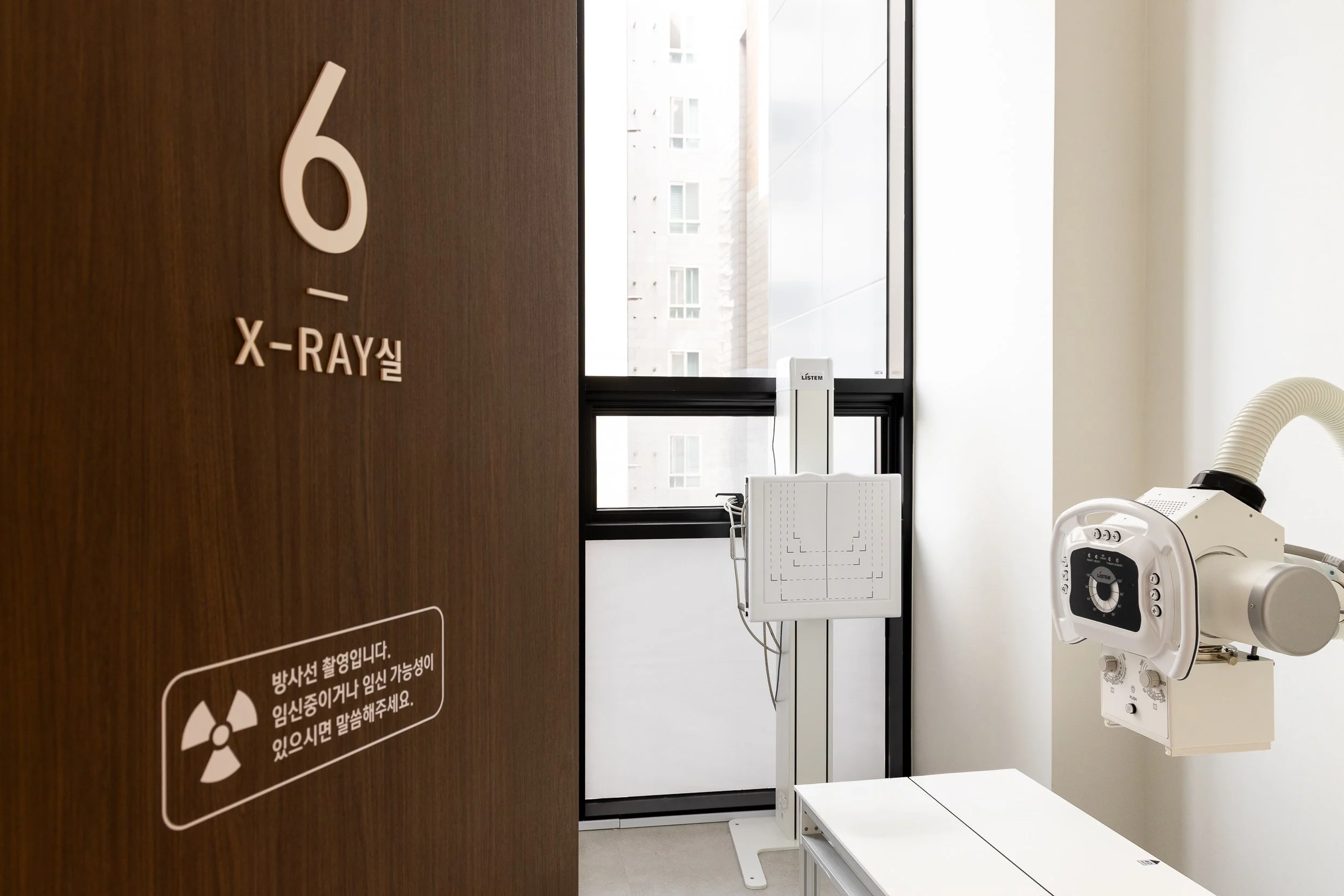 A hospital room with an X-ray machine and a door labeled '6' in large numbers, with some Korean text, and a window in the background.