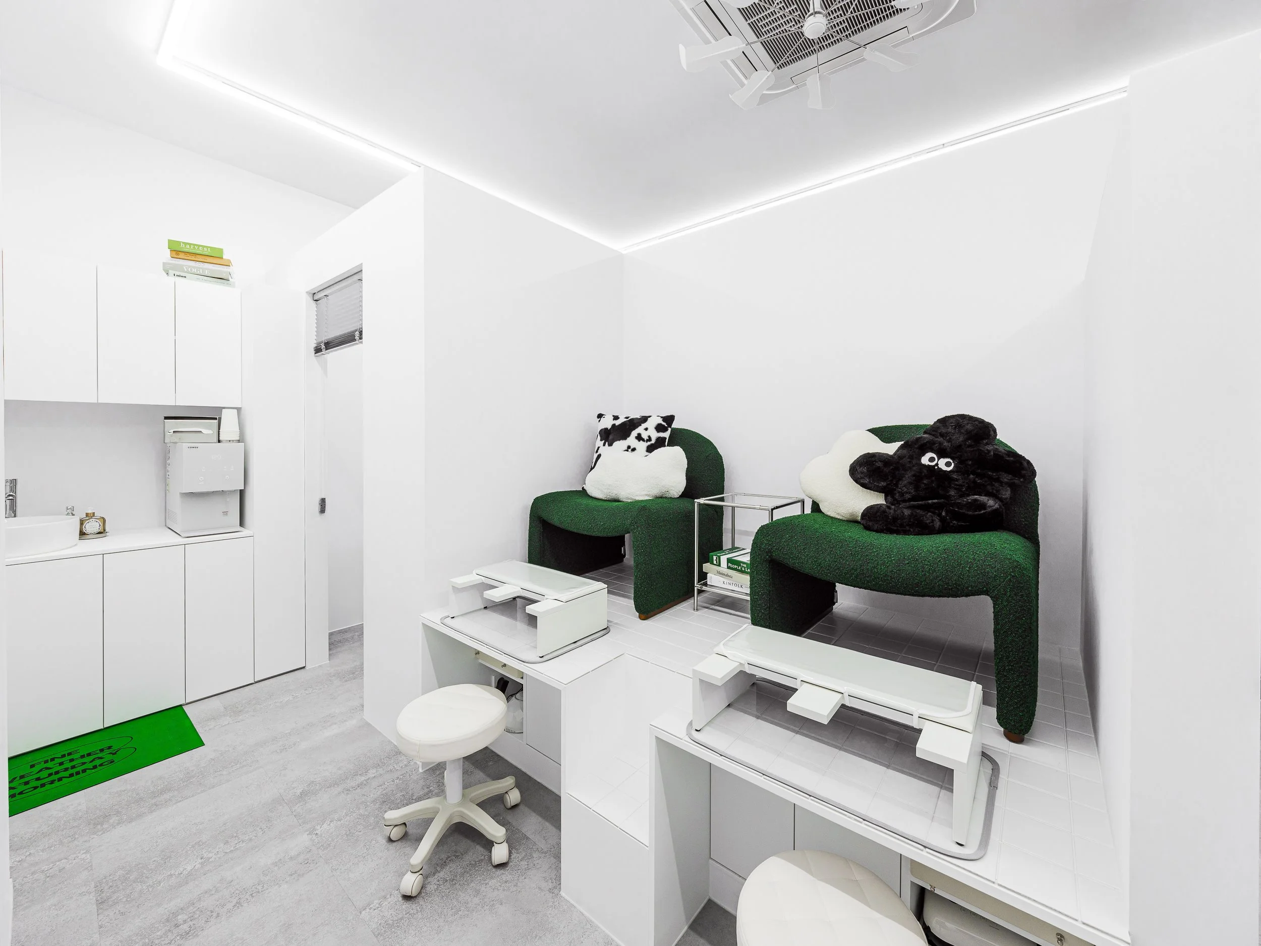 A veterinary clinic examination room with two elevated green chairs with plush pillows, one resembling a cow and the other a dog, on the right side. Medical equipment and supplies, including a small sink, a water dispenser, and storage cabinets, are 