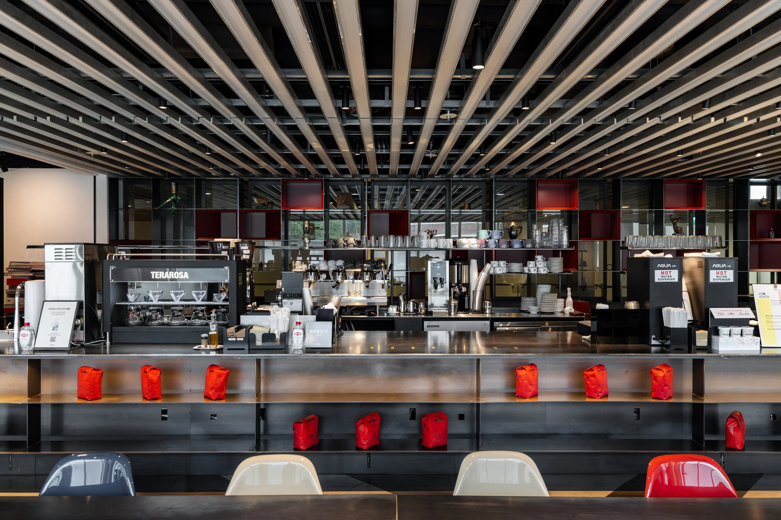 A modern cafe counter with coffee machines, cups, and red bags on the counter, colorful chairs in front, and a modern ceiling design.