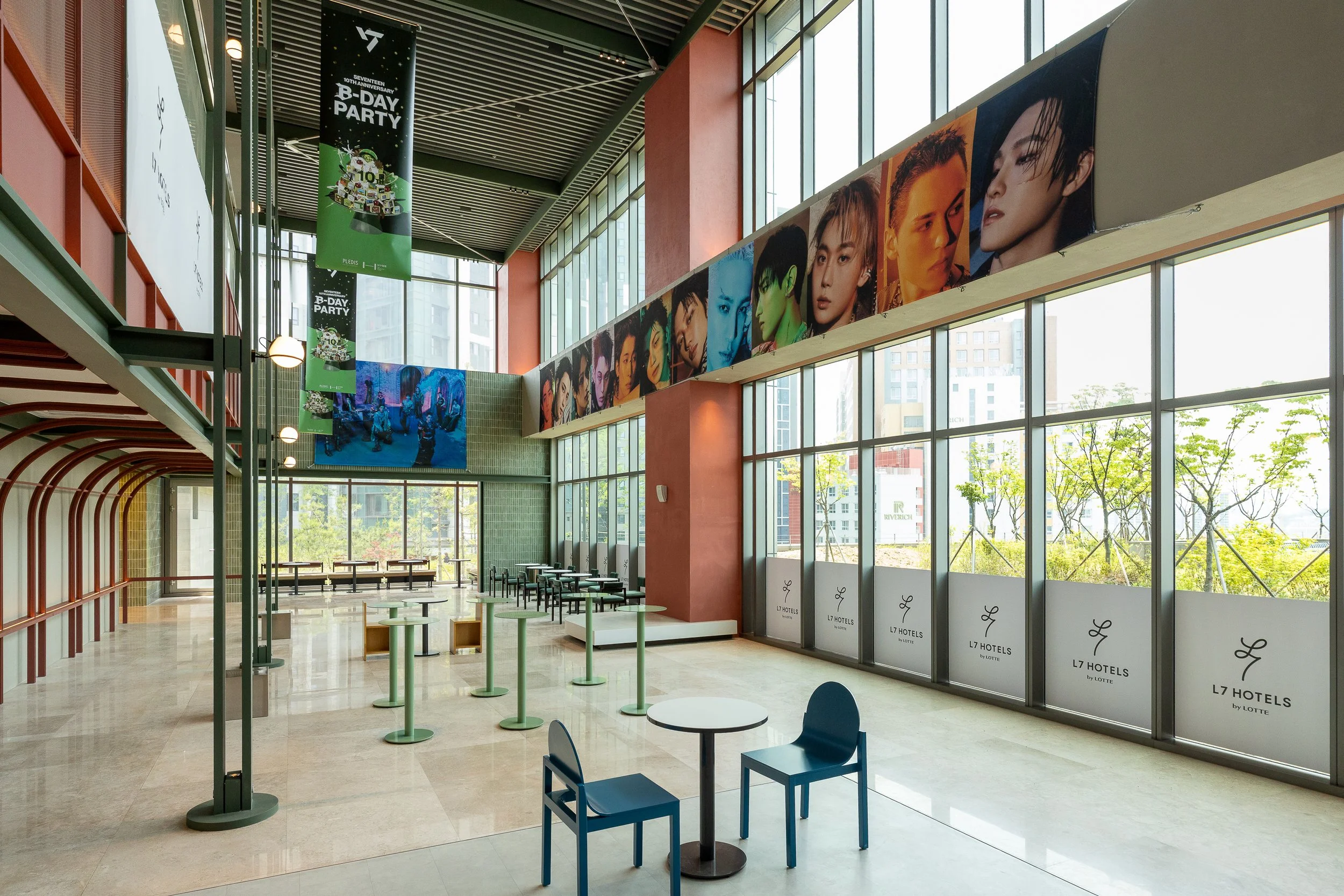 Interior of a hotel lobby with large windows, colorful posters of people on the walls, small tables, and chairs, with some promotional banners hanging from the ceiling.