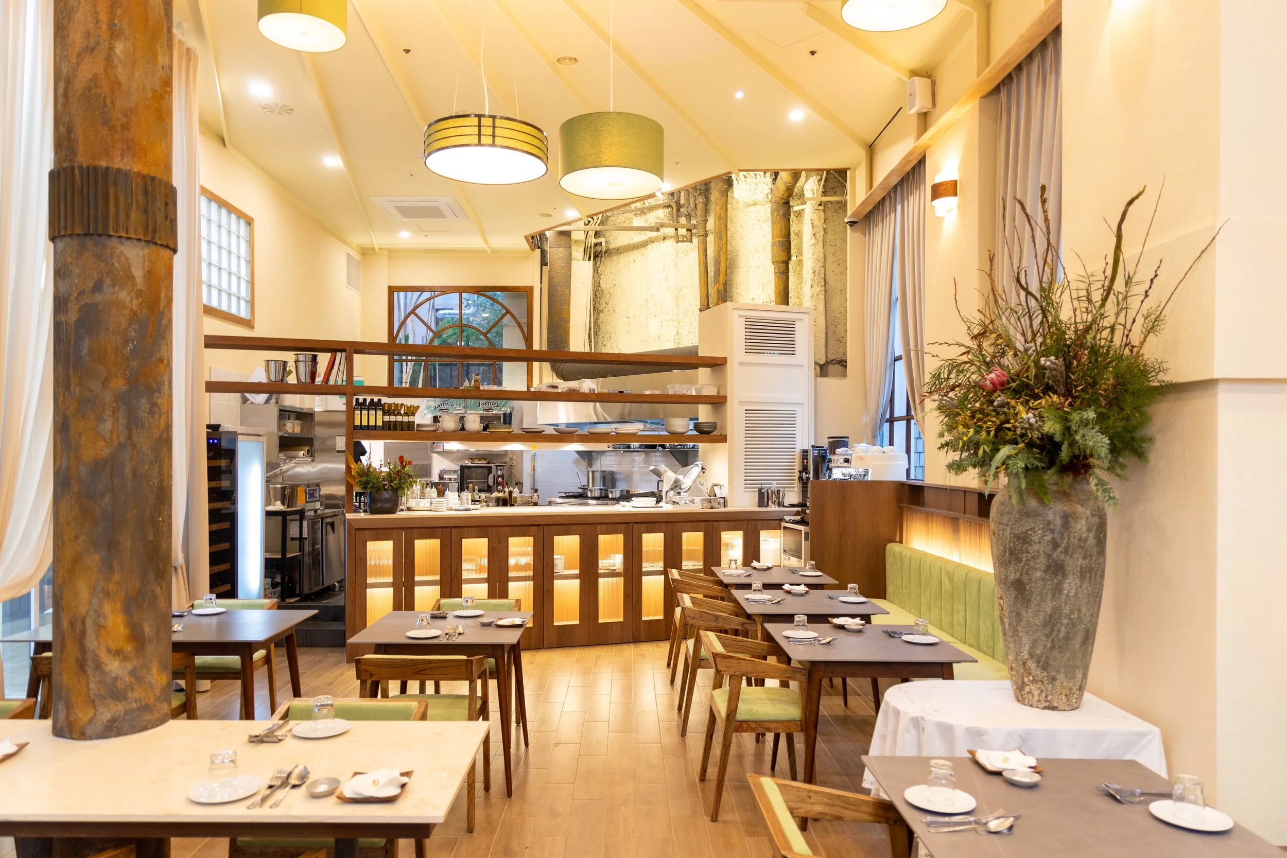 Interior of a restaurant with wooden tables, chairs with green cushions, a large vase of flowers, a bar area with kitchen equipment, and decorative lighting fixtures.
