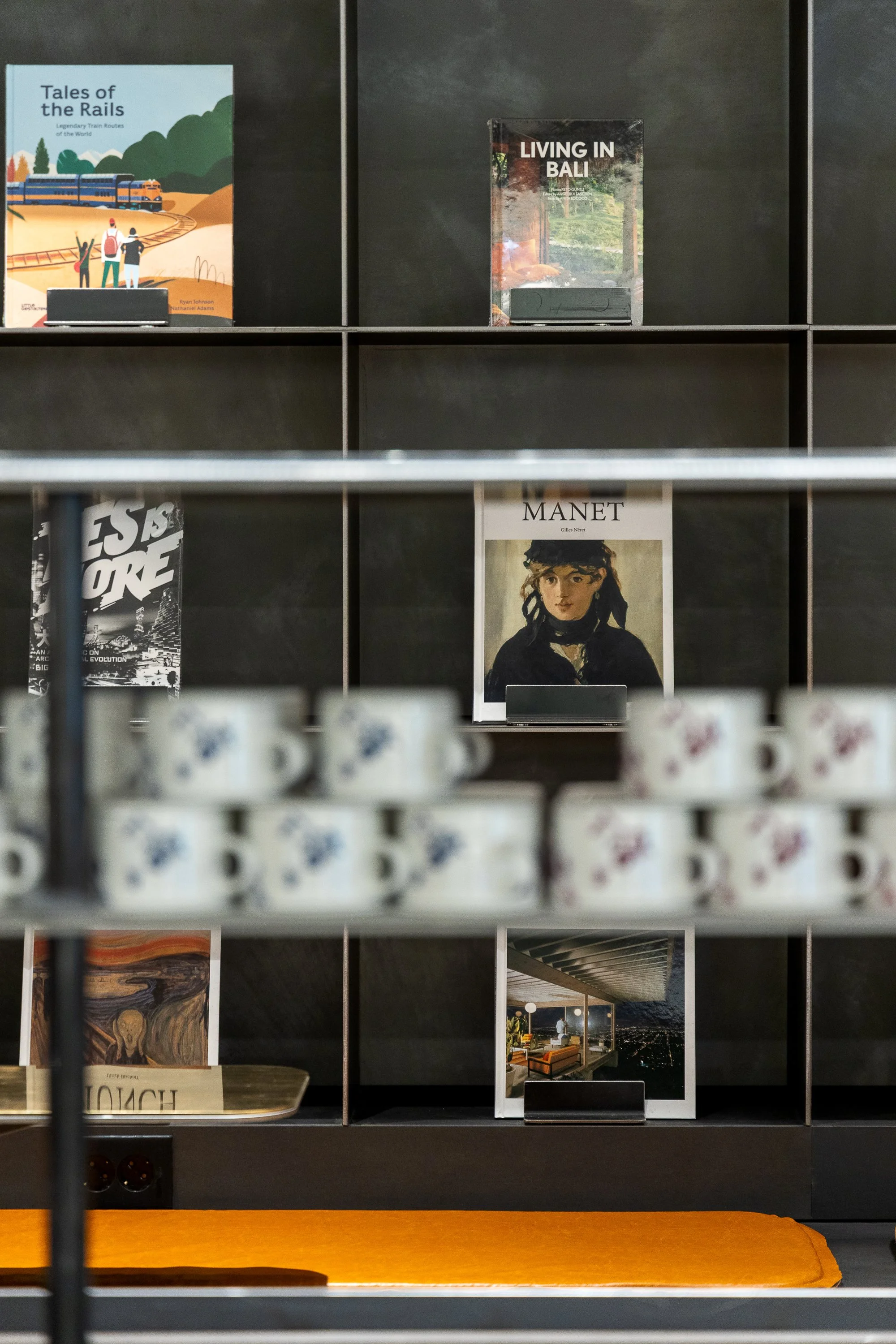 Books and magazines displayed on black shelving; titles include 'Tales of the Rails,' 'Living in Bali,' and 'Manet'; cups with floral designs on a shelf below; orange cushion on a bench in front.