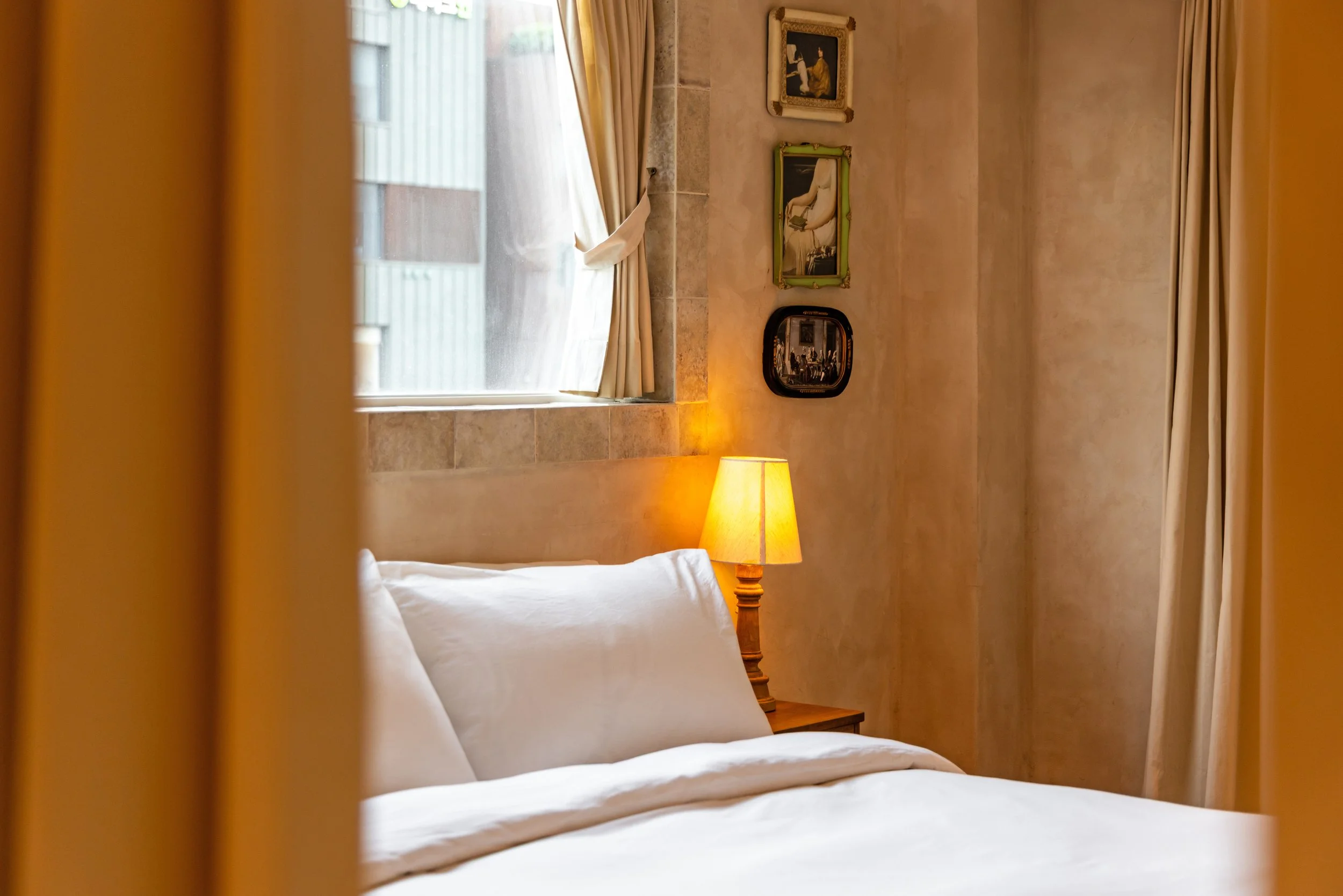 Hotel room with a bed, a nightstand with a lamp, a window with curtains, and wall art.