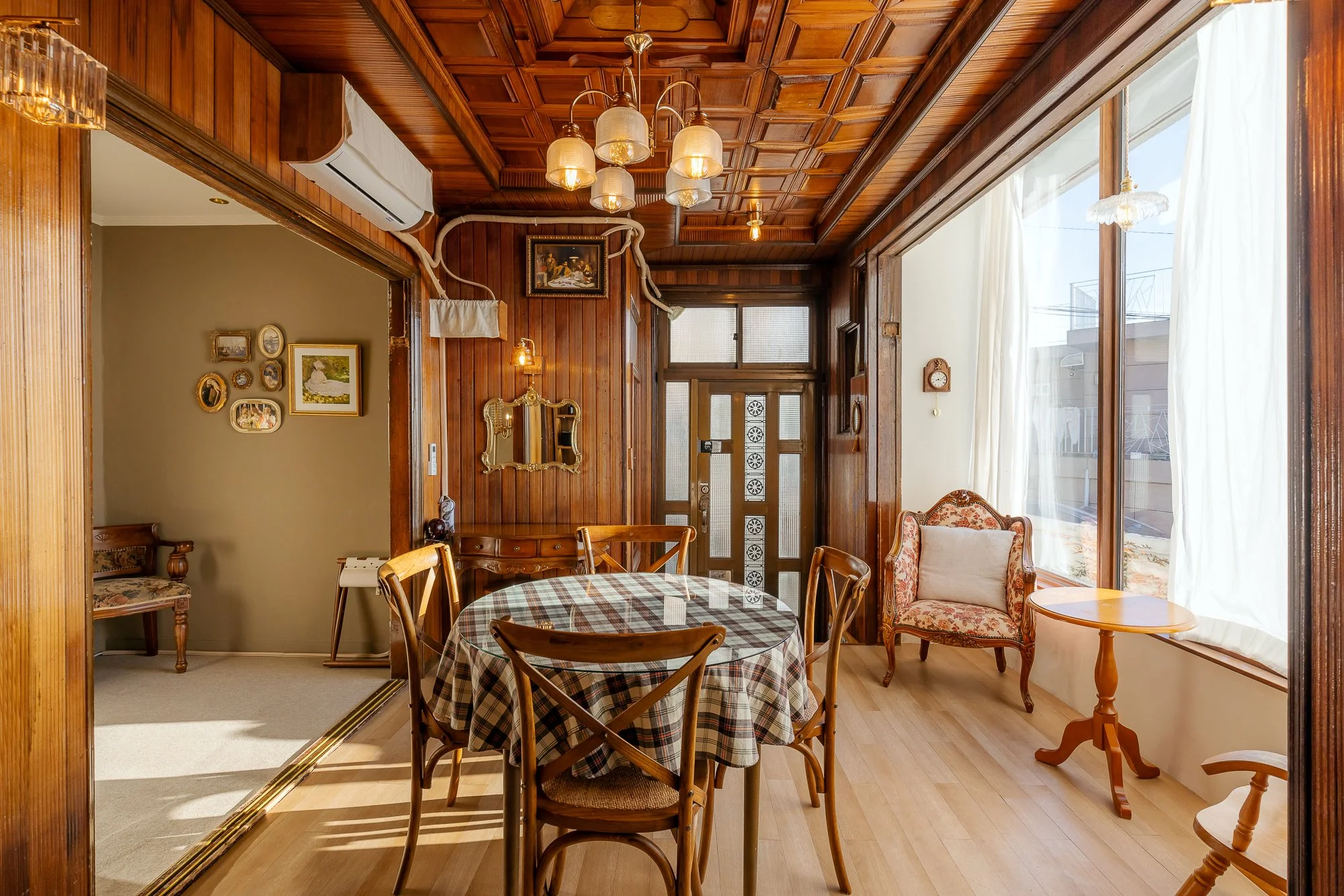 A cozy dining room with wooden walls and ceiling, a round dining table with a checkered tablecloth, six matching chairs, vintage armchair, small side tables, large bay window with white curtains, and decorative wall art.