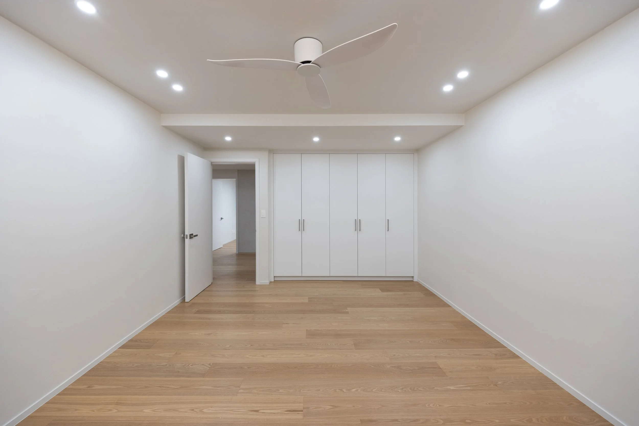 Empty room with white walls, wooden floor, ceiling fan, recessed lighting, open door, and built-in white closet.