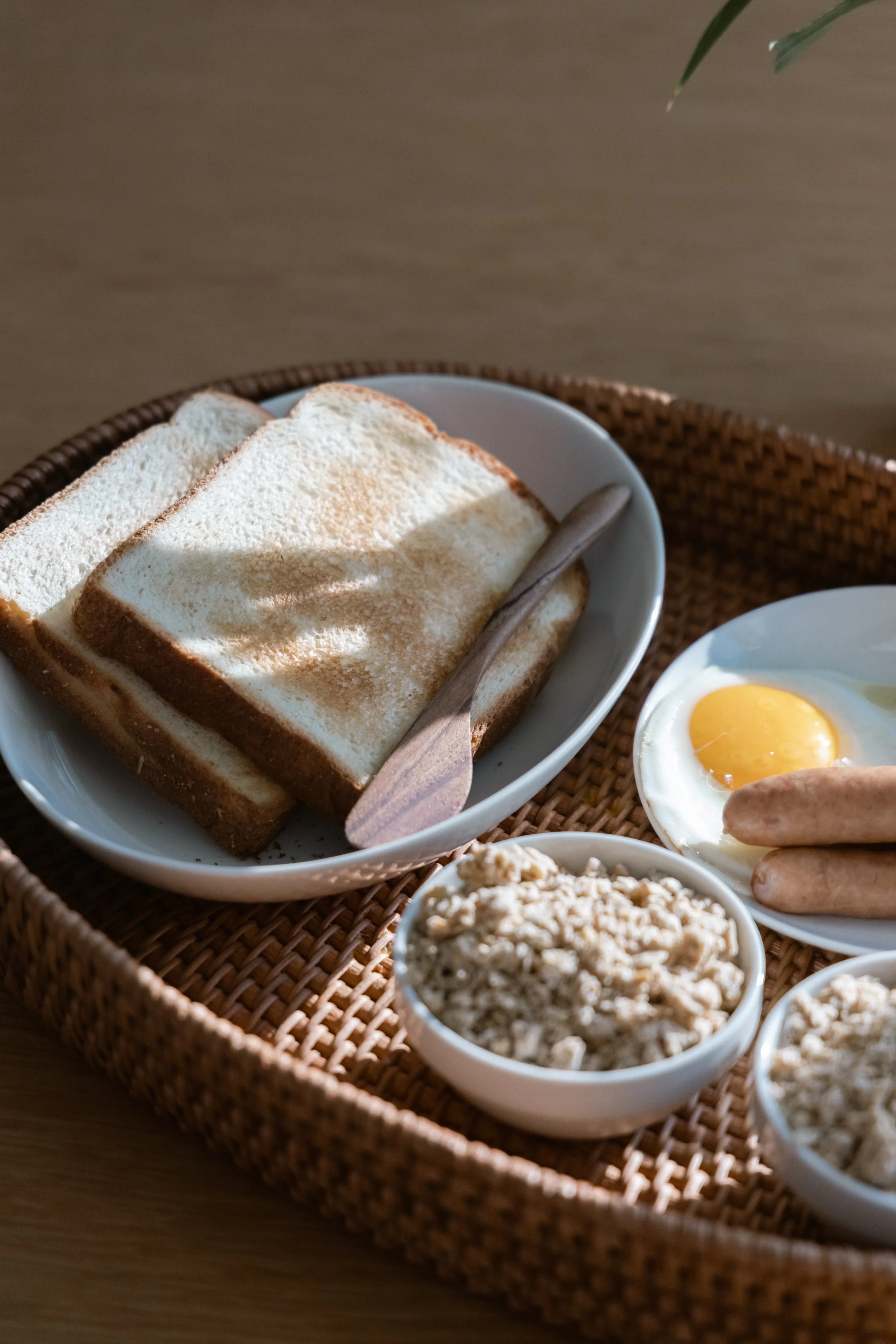 Tray with sliced bread, a fried egg, sausages, and bowls of cereal on a wooden surface.