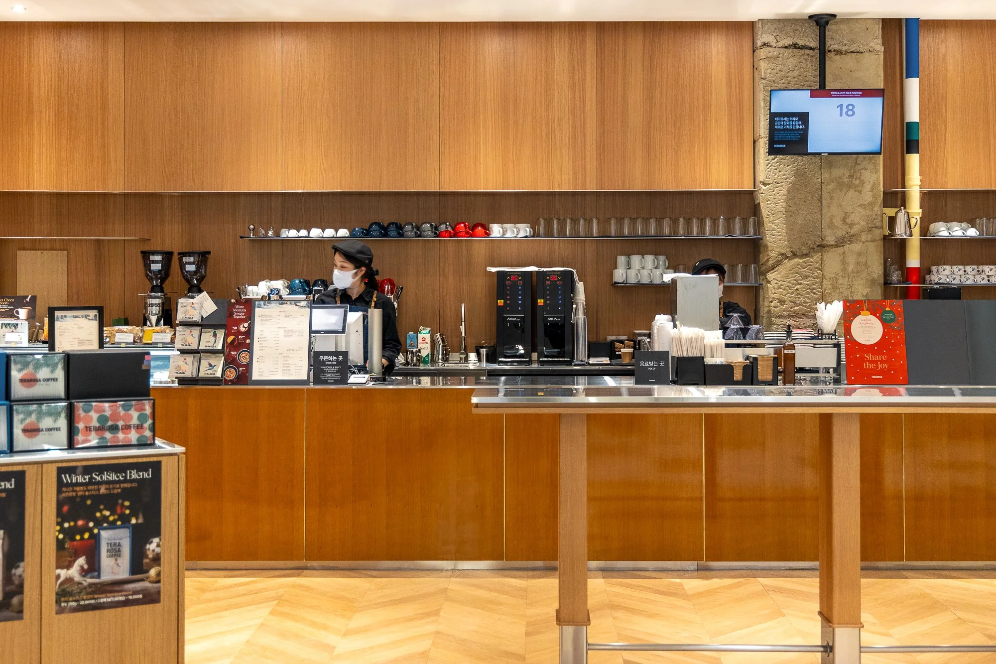 A coffee shop counter with two baristas wearing masks, wooden wall background, shelves with cups and accessories, coffee machines, a digital screen, and a red holiday-themed sign.