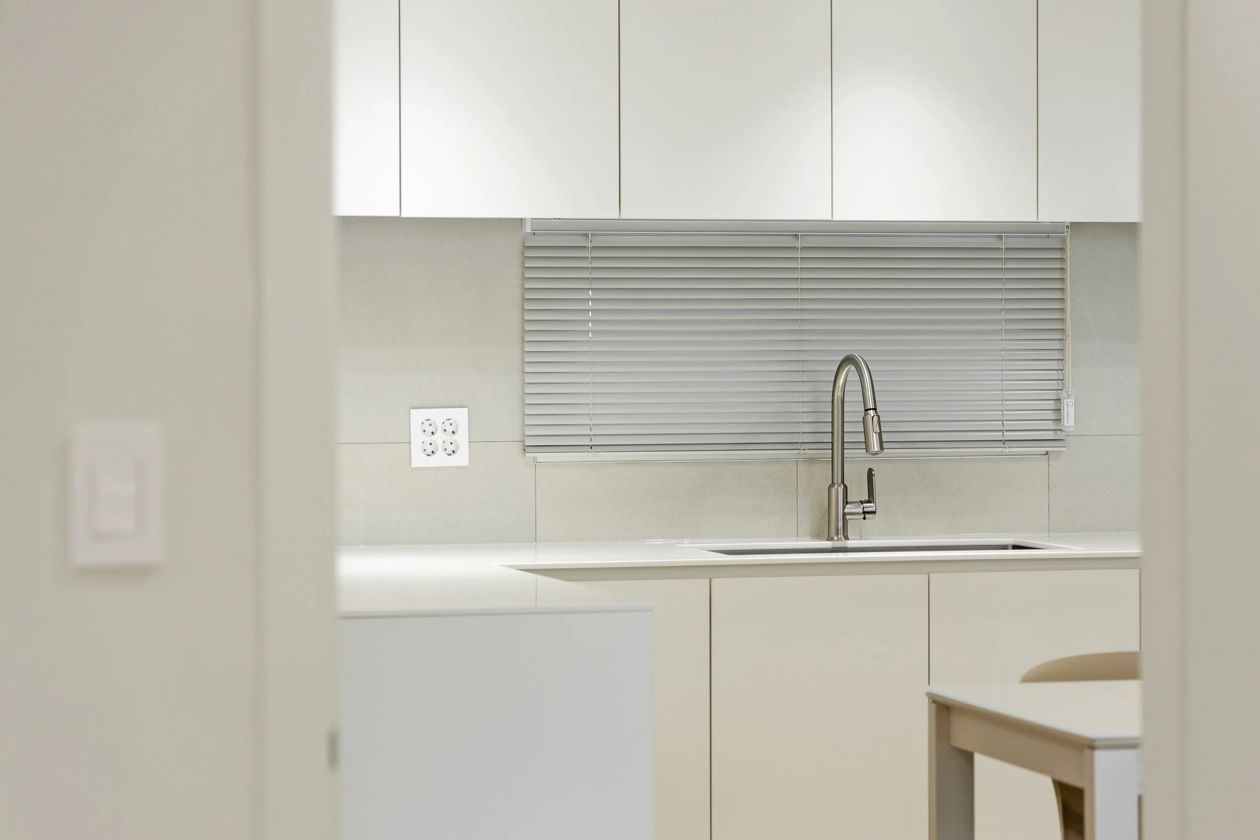 A minimalist kitchen with a stainless steel faucet, white cabinets, and a window with closed horizontal blinds.