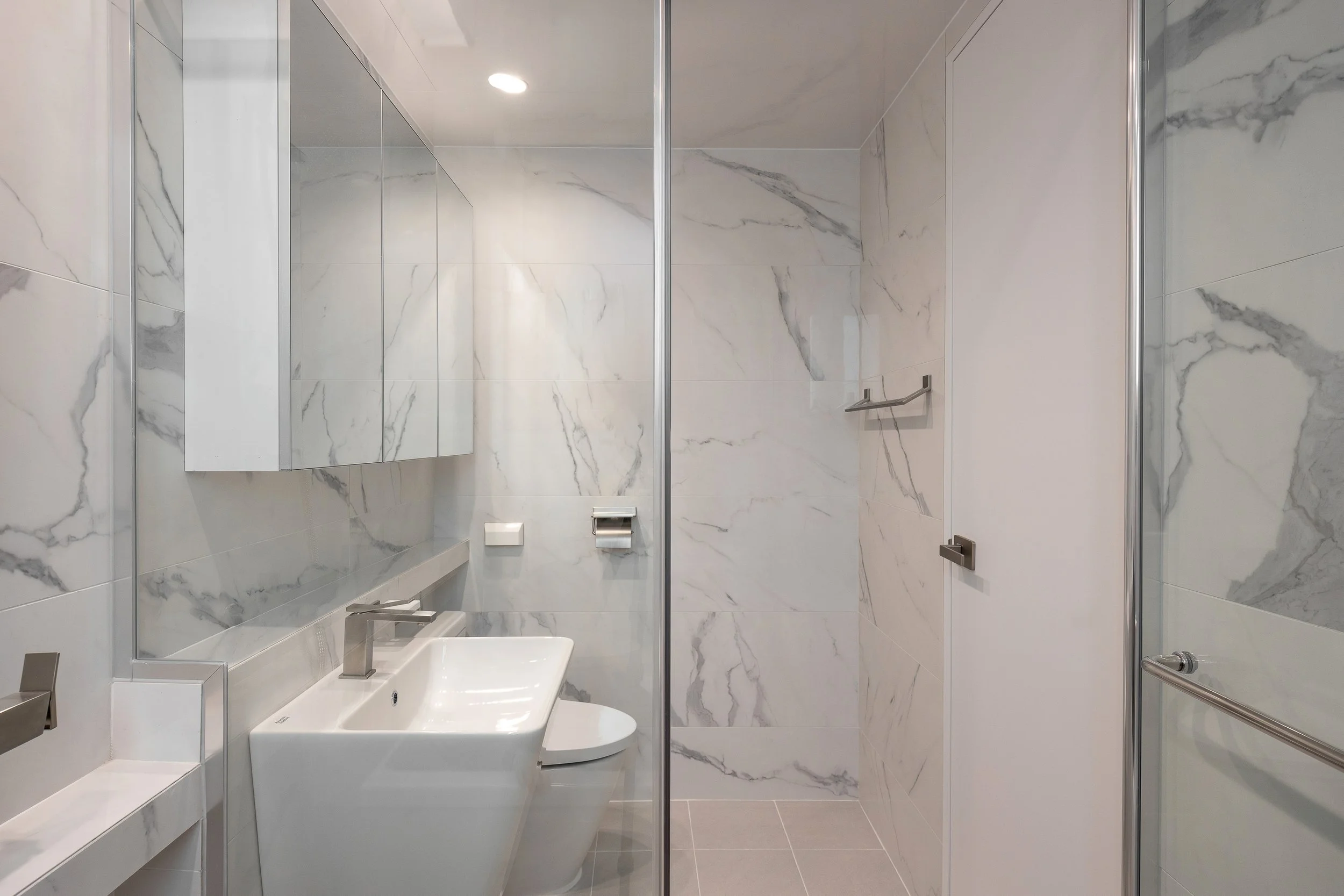 Modern bathroom with white marble walls, a white sink, a mirror cabinet, a toilet, a towel bar, and a glass shower door.