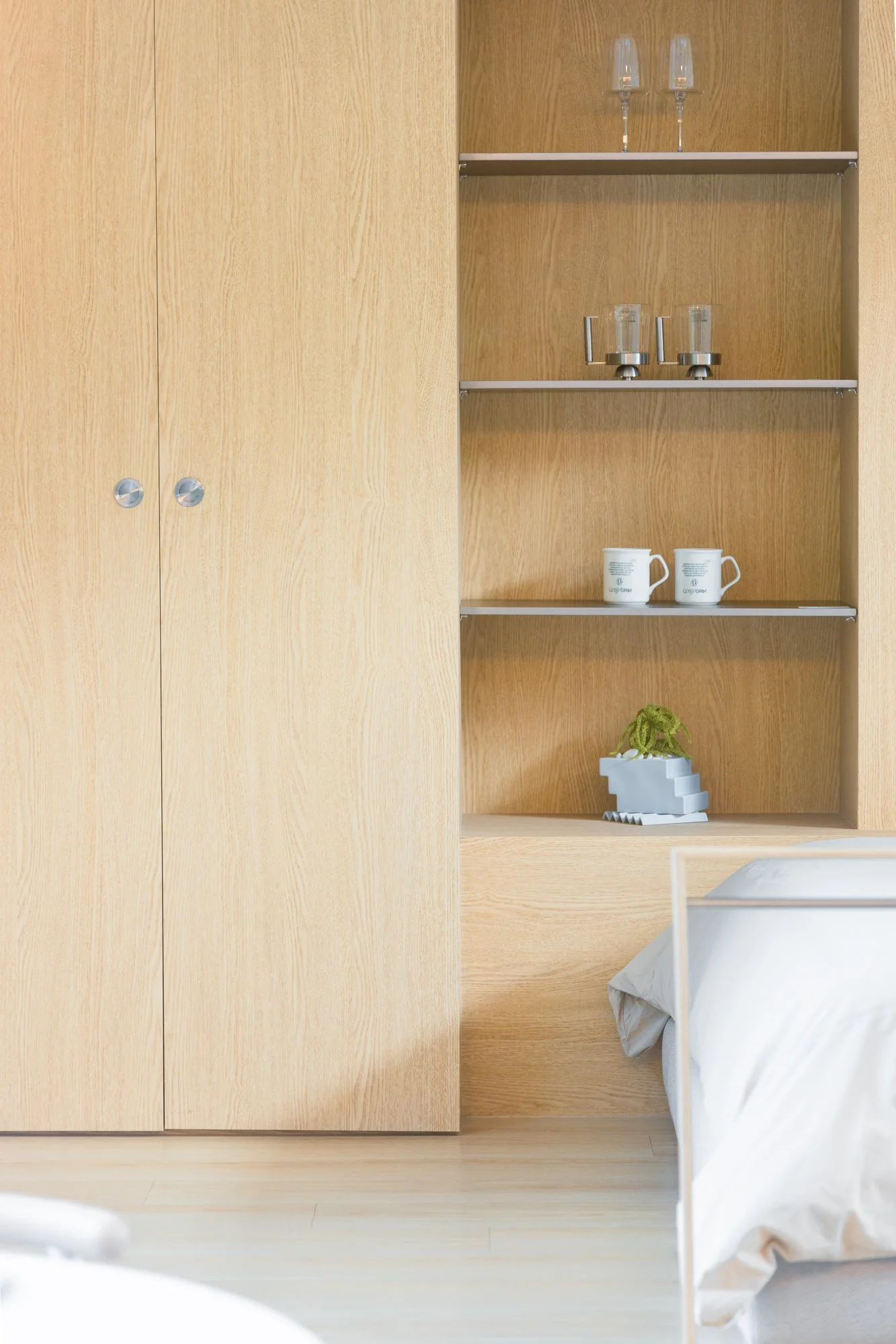 Interior of a bedroom with wooden built-in wardrobe and open shelving, holding glasses, mugs, and a small plant. Part of a bed with white bedding is visible.
