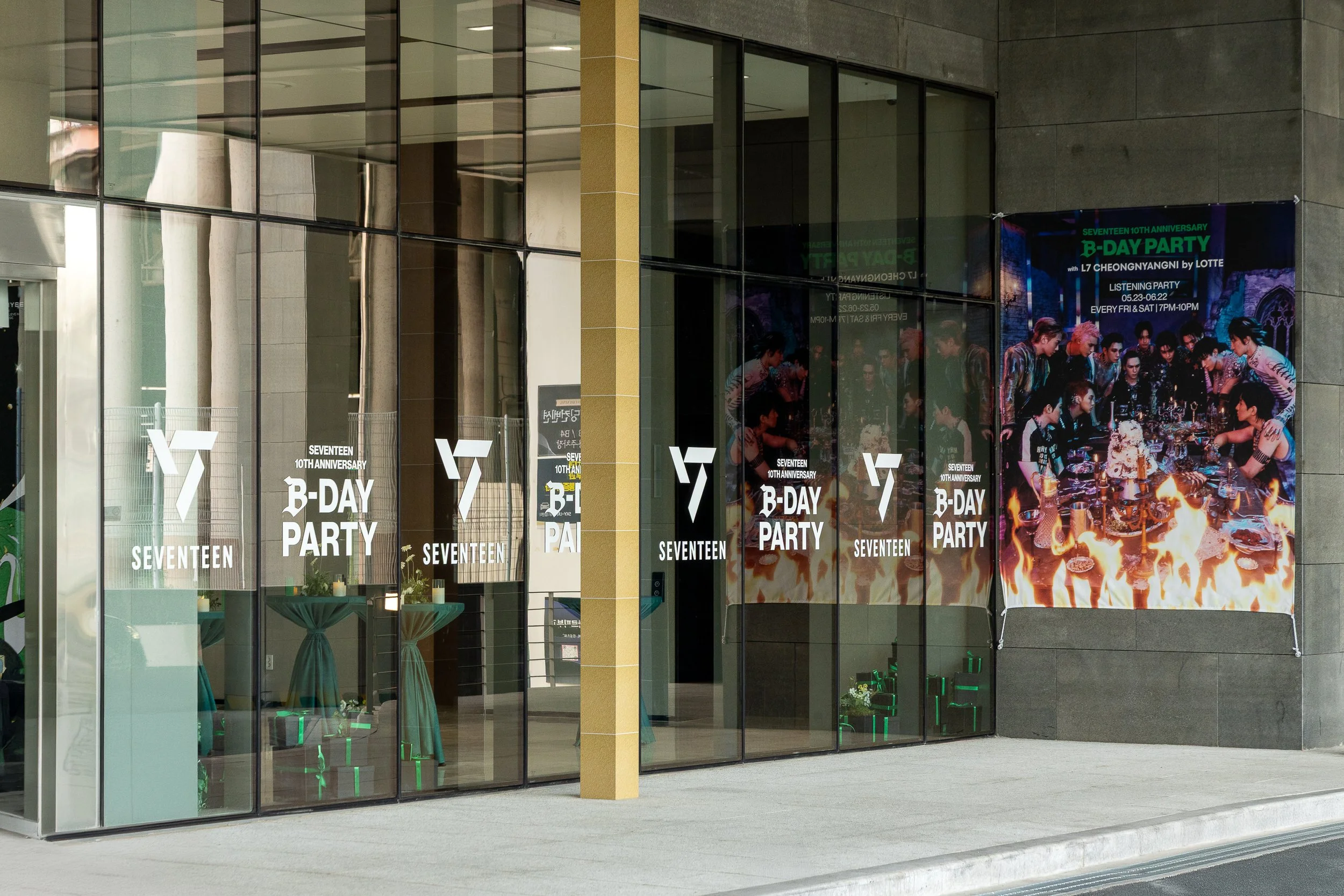 Glass building windows with banners and posters advertising SEVENTEEN's 10th anniversary B-day party, featuring a large group of people celebrating with fire and food.
