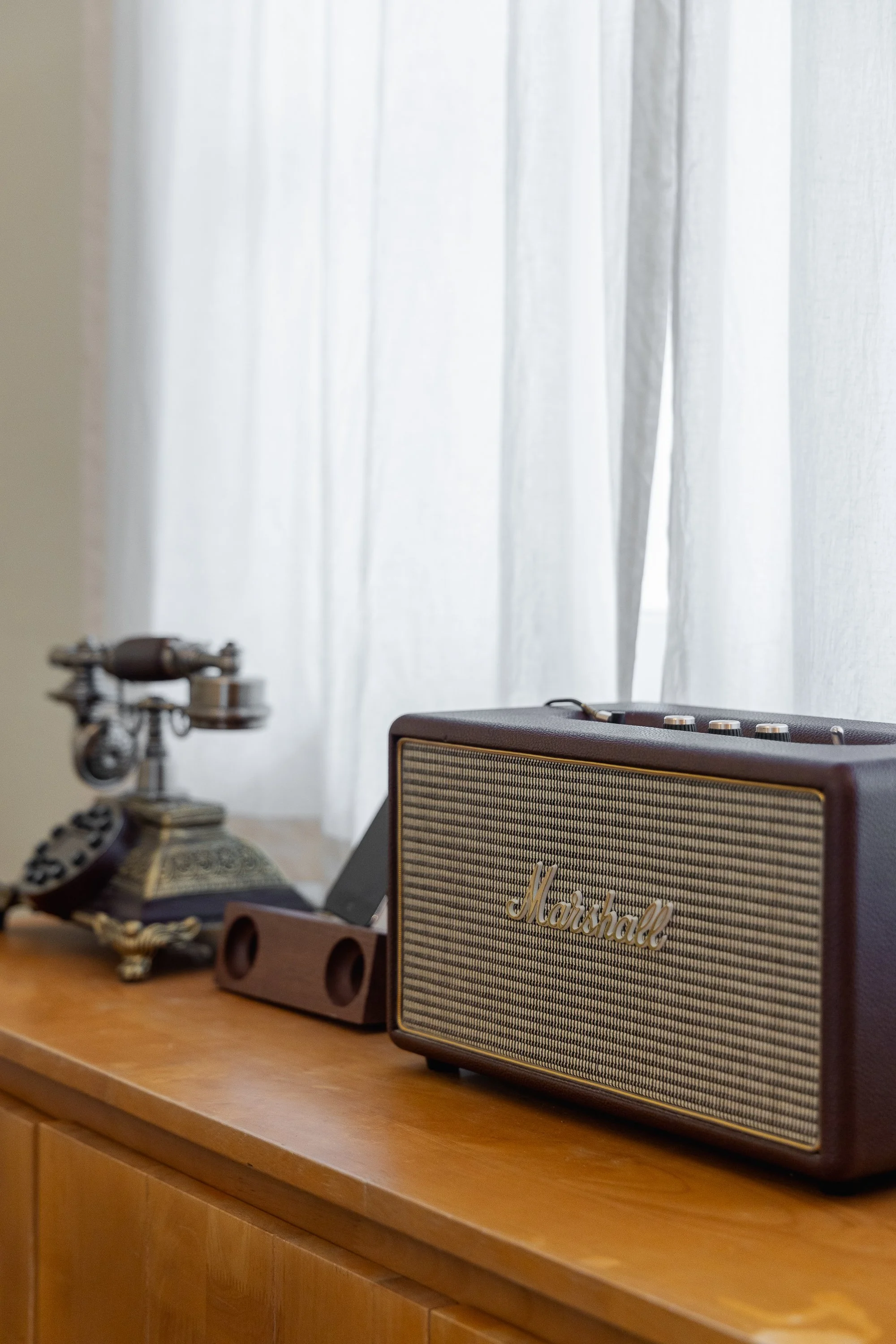 Close-up of a vintage rotary dial telephone, a Marshall speaker, and a wooden speaker stand sitting on a wooden surface with a white curtain in the background.