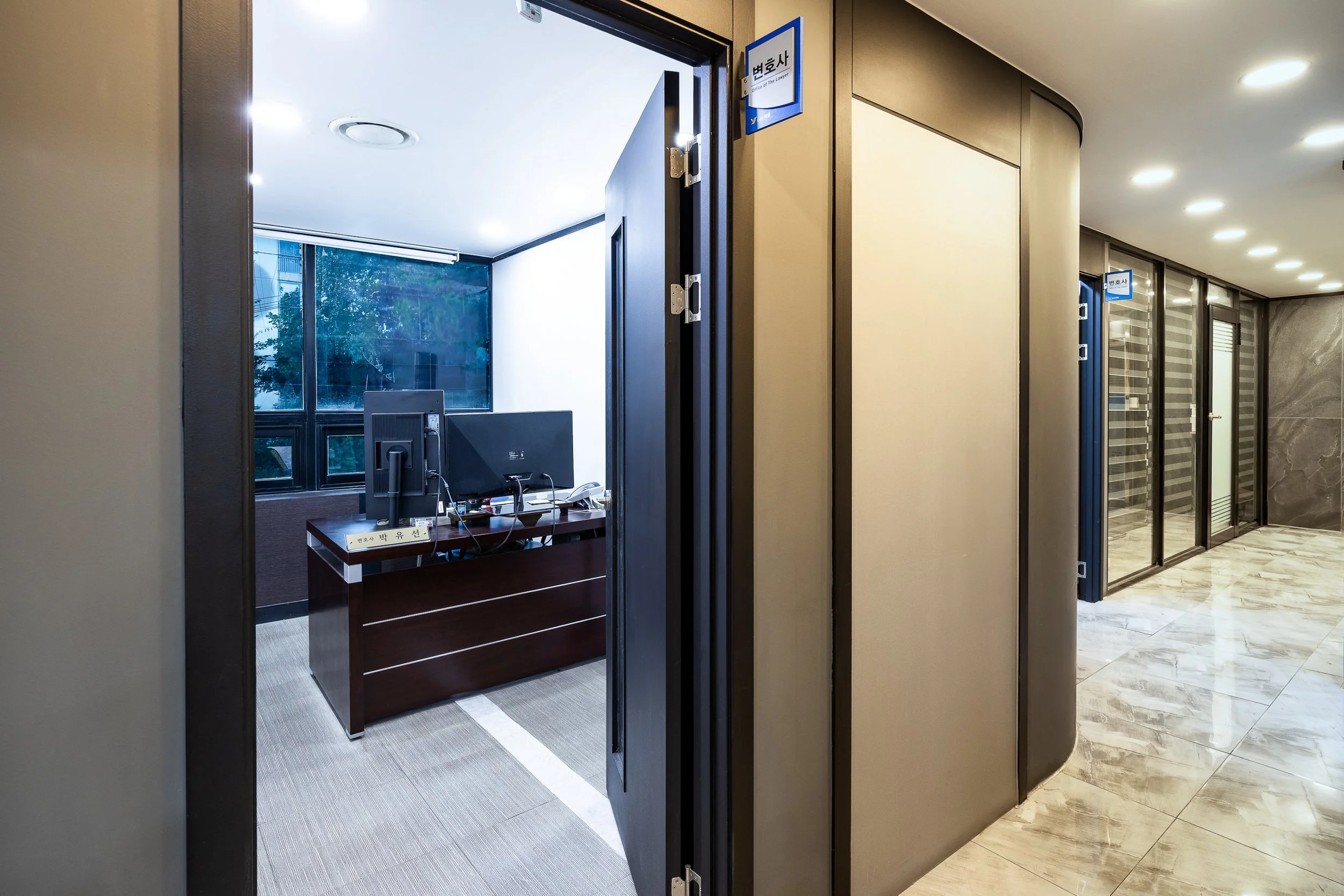Office reception area with a desk, computer, and a large window, adjacent to a hallway with glass doors and marble flooring.