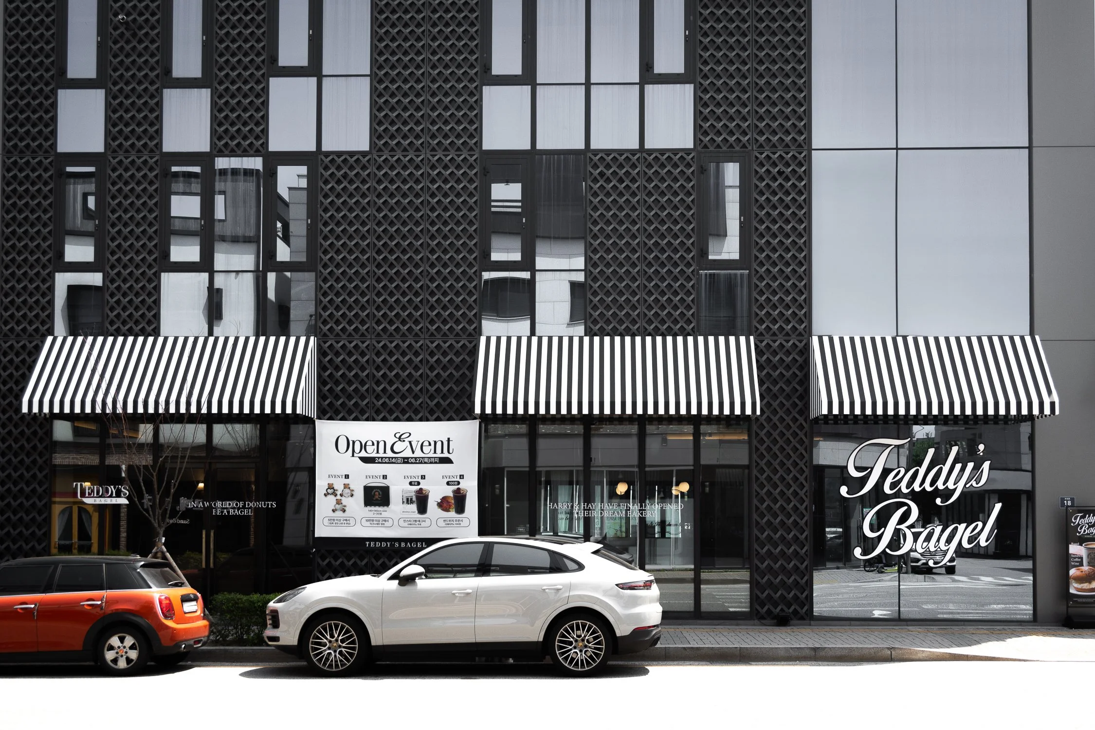 Front view of a modern building with black lattice and window structure, two striped black and white awnings, and signs for "Teddy's Bagel" and "Open & Event," with parked white and orange cars in front.