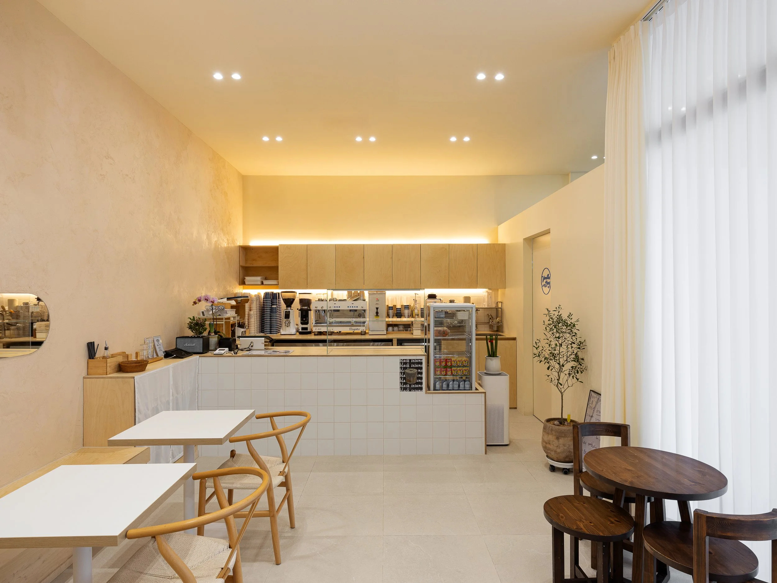 Bright, minimalist cafe interior with white tables and wooden chairs, a coffee counter with equipment, potted plants, and large windows with white curtains.