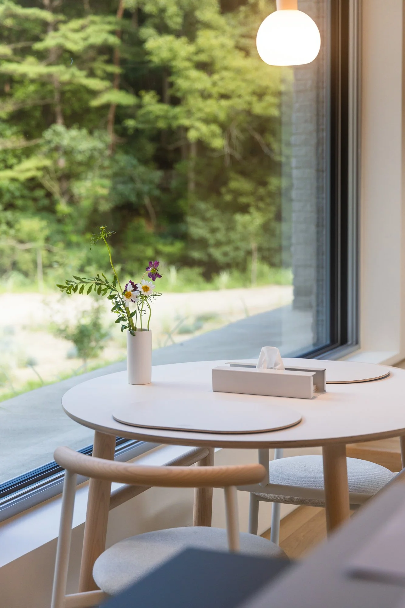 A round white dining table with a small white vase holding wildflowers, a box of tissues, and placemats, positioned near a large window overlooking a green forest.