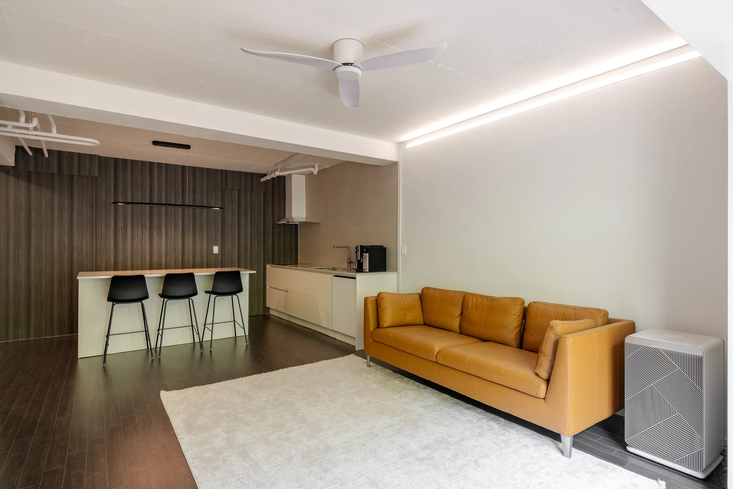 Modern living room and kitchen area with a tan leather sofa, white area rug, dark hardwood floors, white wall, kitchen counter with three black barstools, wooden accent wall, ceiling fan, and sleek lighting.
