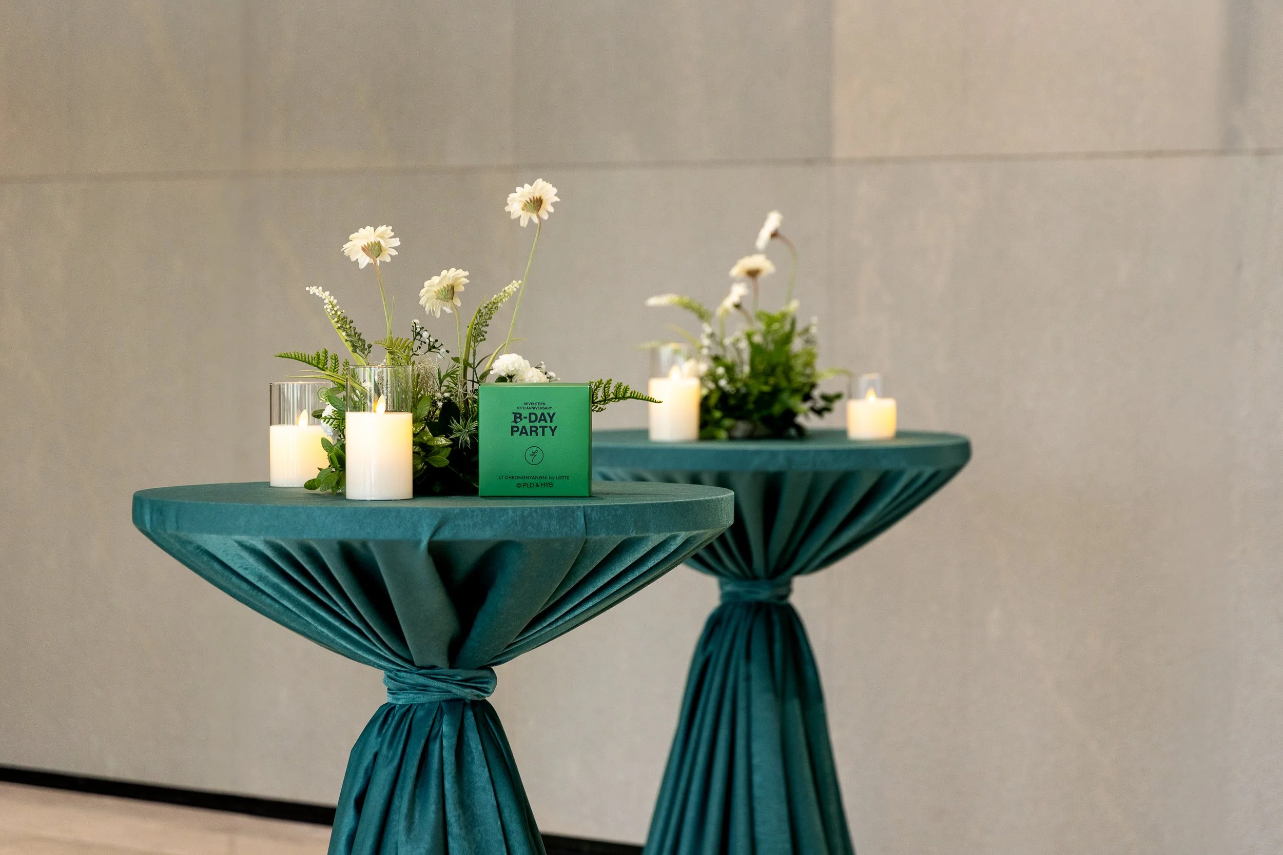 Two round tables draped in teal cloth with pleated skirts tied in the middle, decorated with white candles and floral arrangements of daisies and greenery, and a green sign that reads 'B-DAY PARTY'.