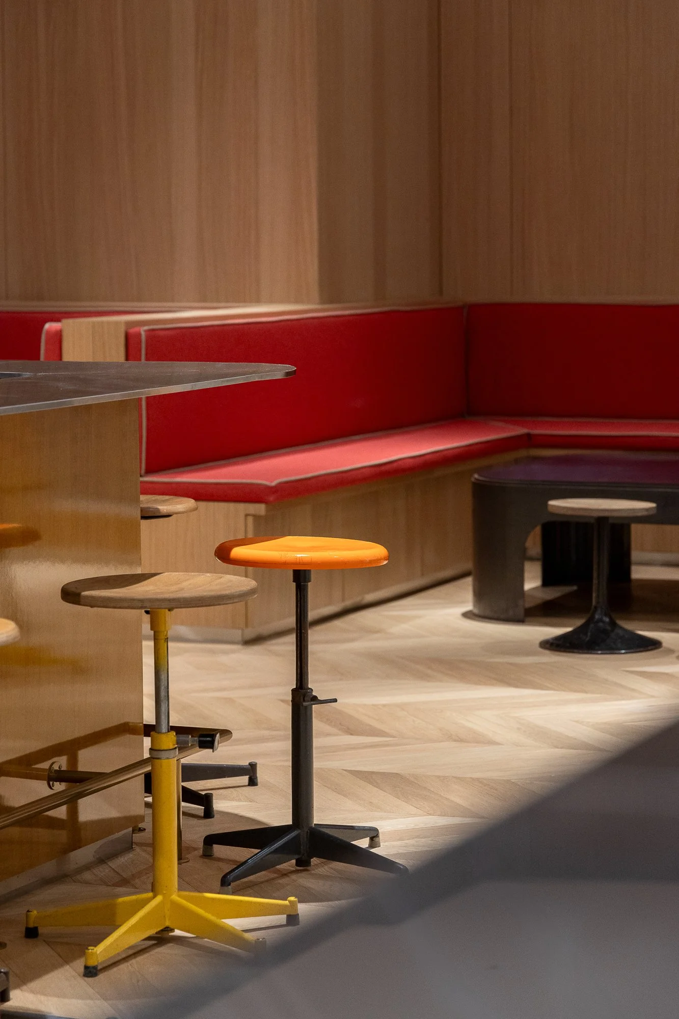 Interior of a cafe with wooden flooring, a red cushioned booth, small round tables, and colorful stools in yellow, black, and orange.