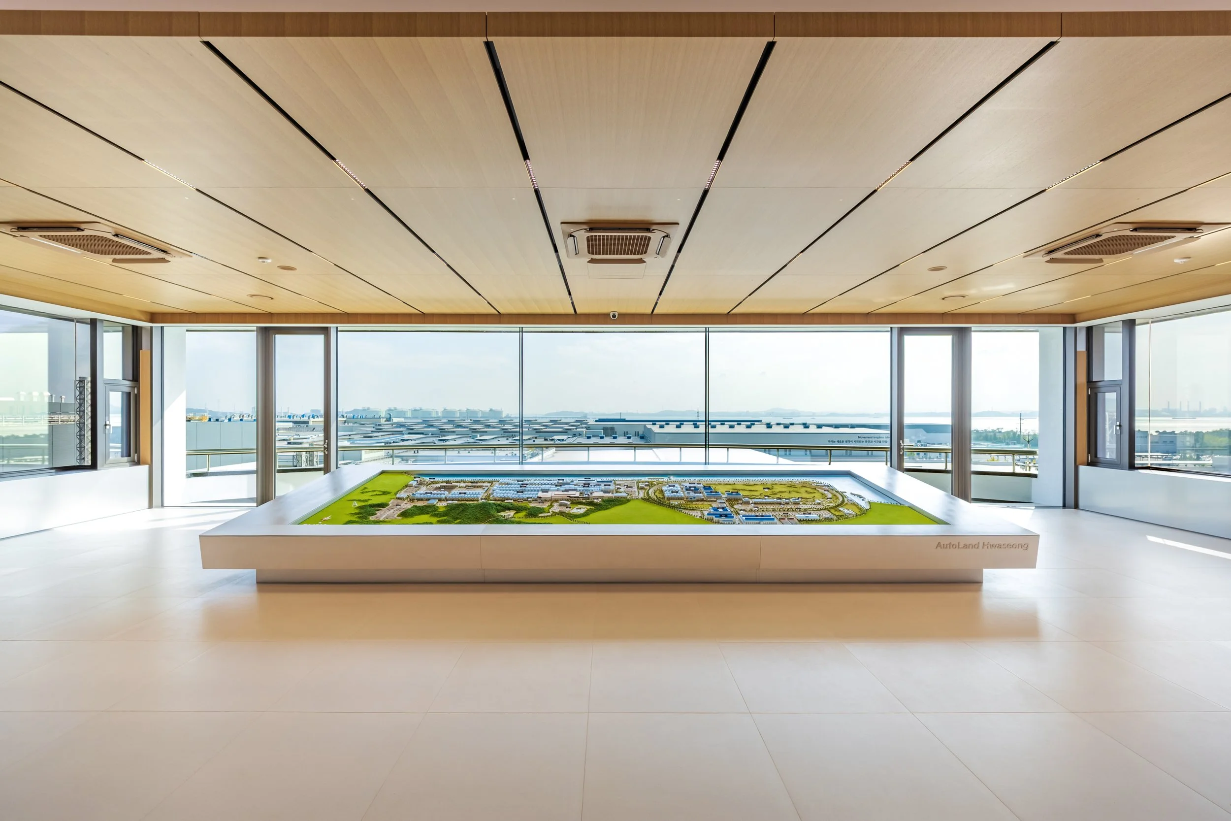 A large indoor room with a detailed scale model of a development project on a white table. The room has a wooden ceiling, white walls, and large windows with city and water views.