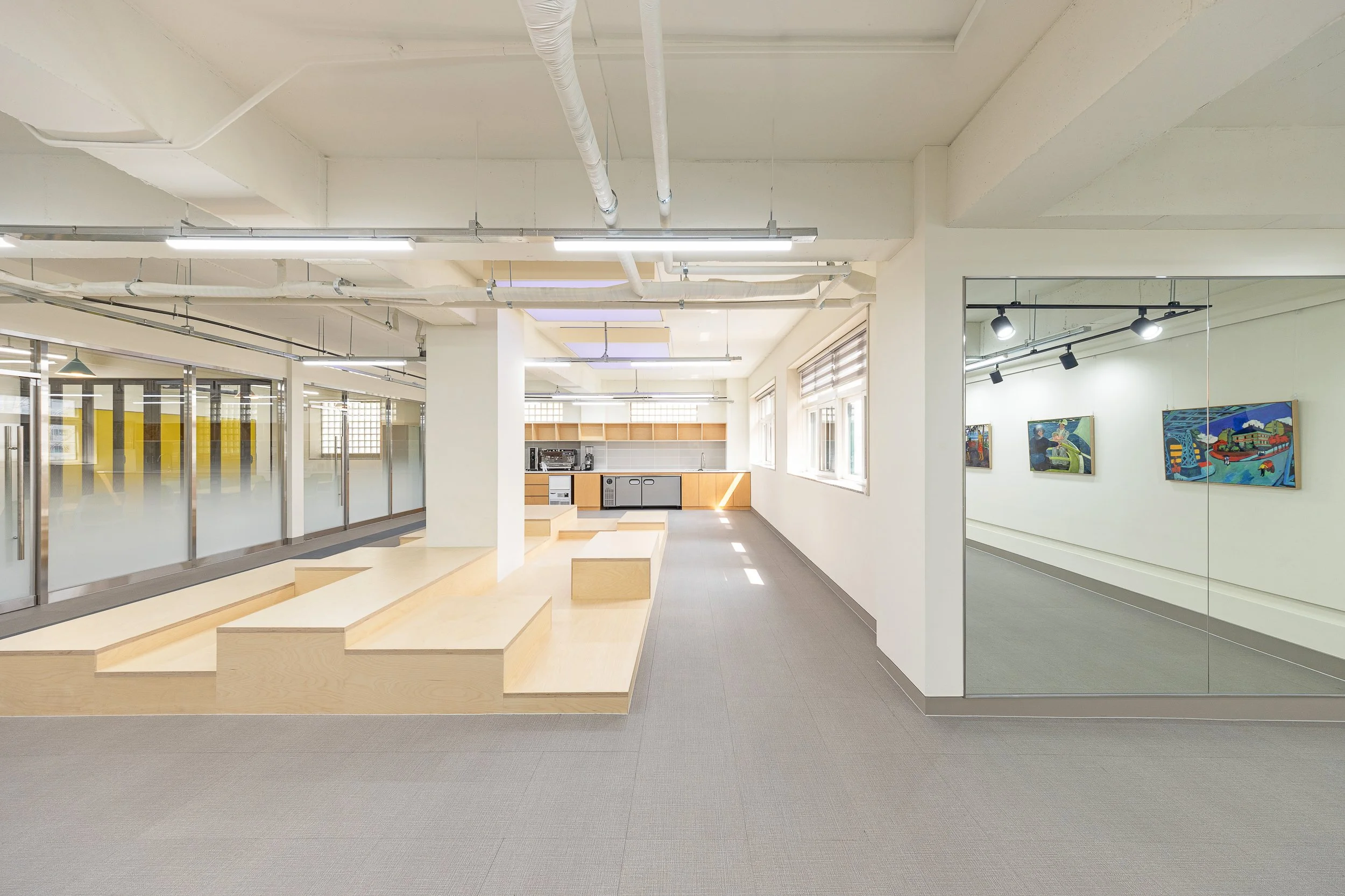 Interior of an art gallery featuring paintings on the wall, a mirrored wall reflecting lights and artworks, a room with large windows allowing natural light, and a seating area with wooden benches.